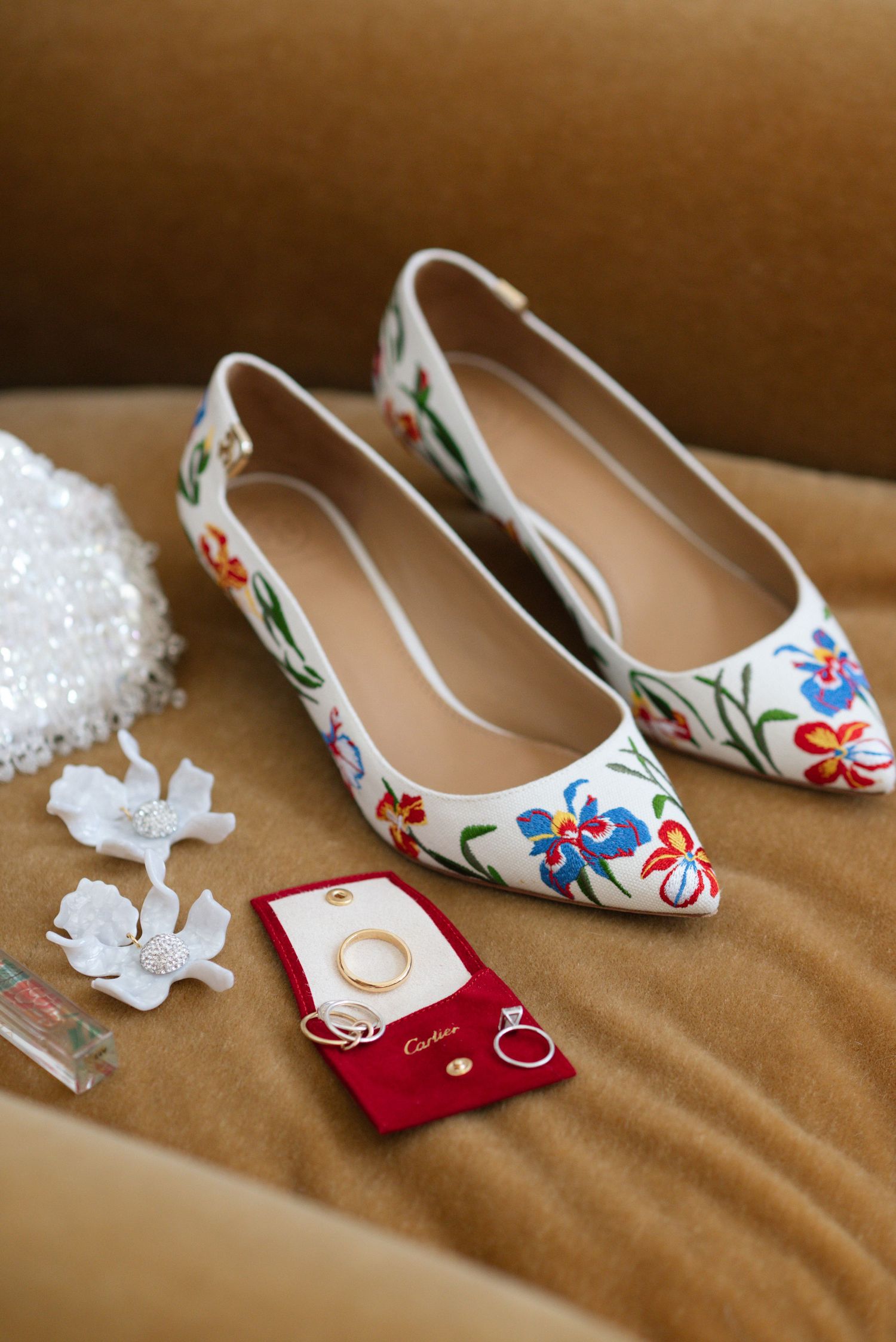 White embroidered bridal shoes with wedding rings and invitation on brown surface.