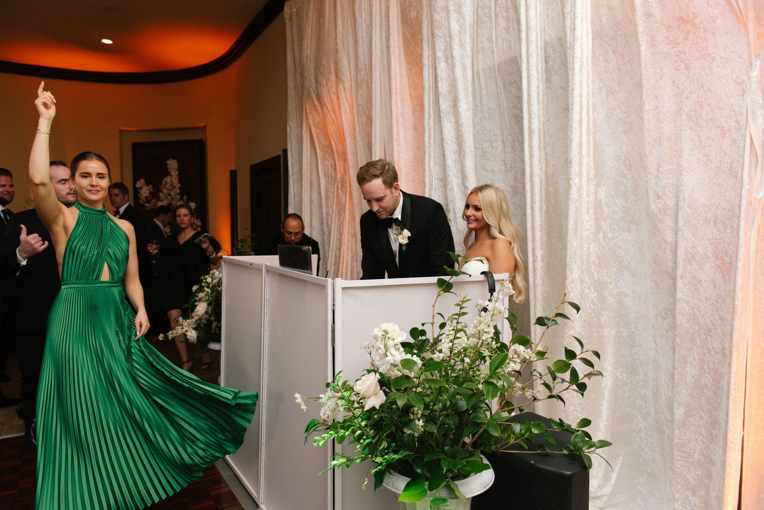 Wedding reception scene with a green pleated dress in focus as guests gather near a decorated white podium.
