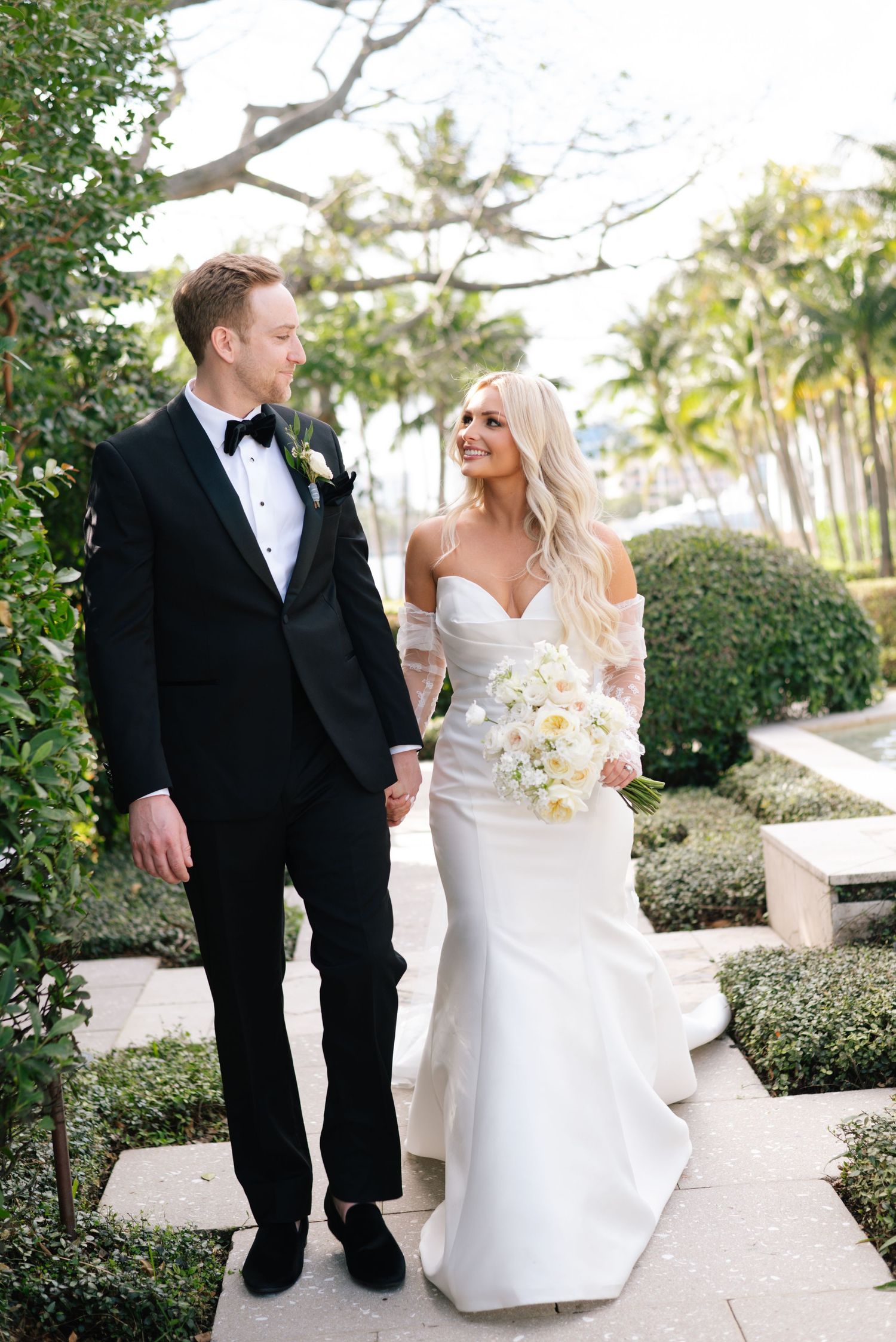 Wedding couple shares intimate moments along a garden path with manicured hedges and palm trees.
