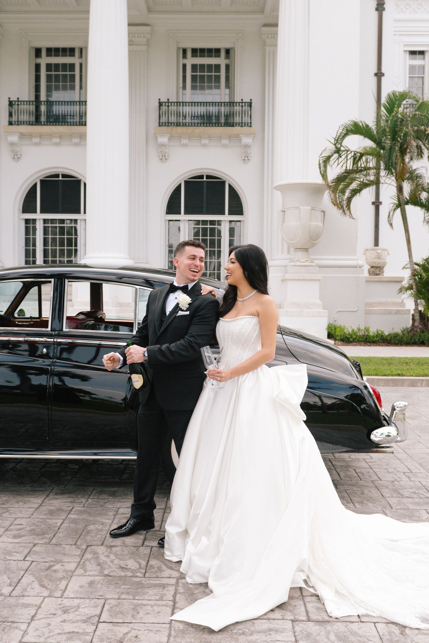 A wedding couple poses playfully in front of a classic white building and vintage car.