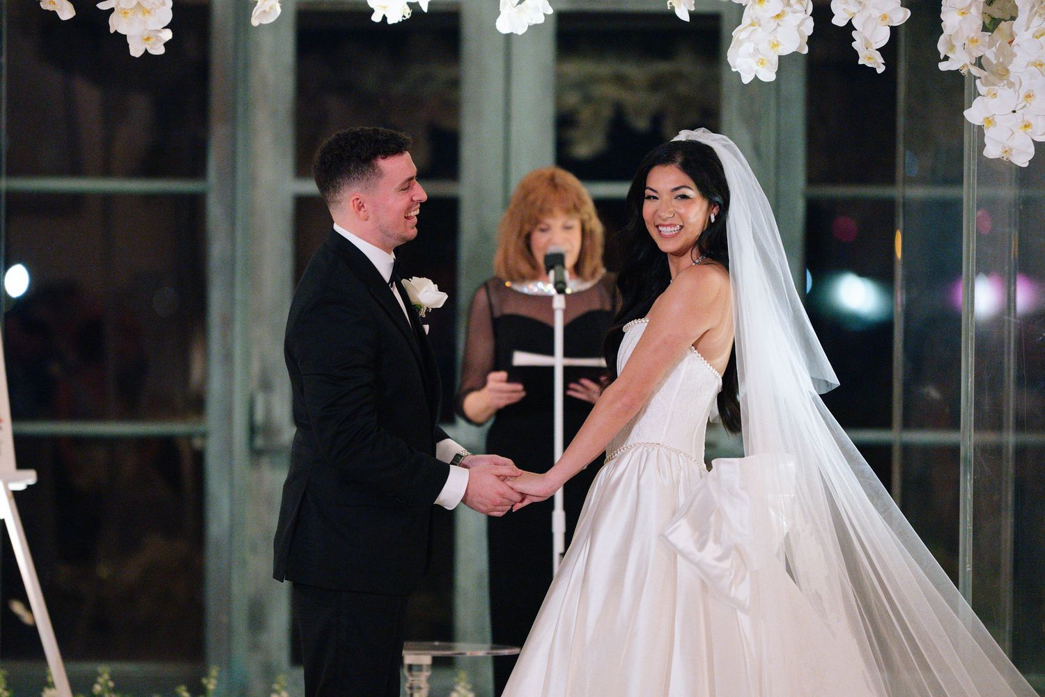 Wedding ceremony in ornate ballroom with hanging white floral arrangements and formal guests.