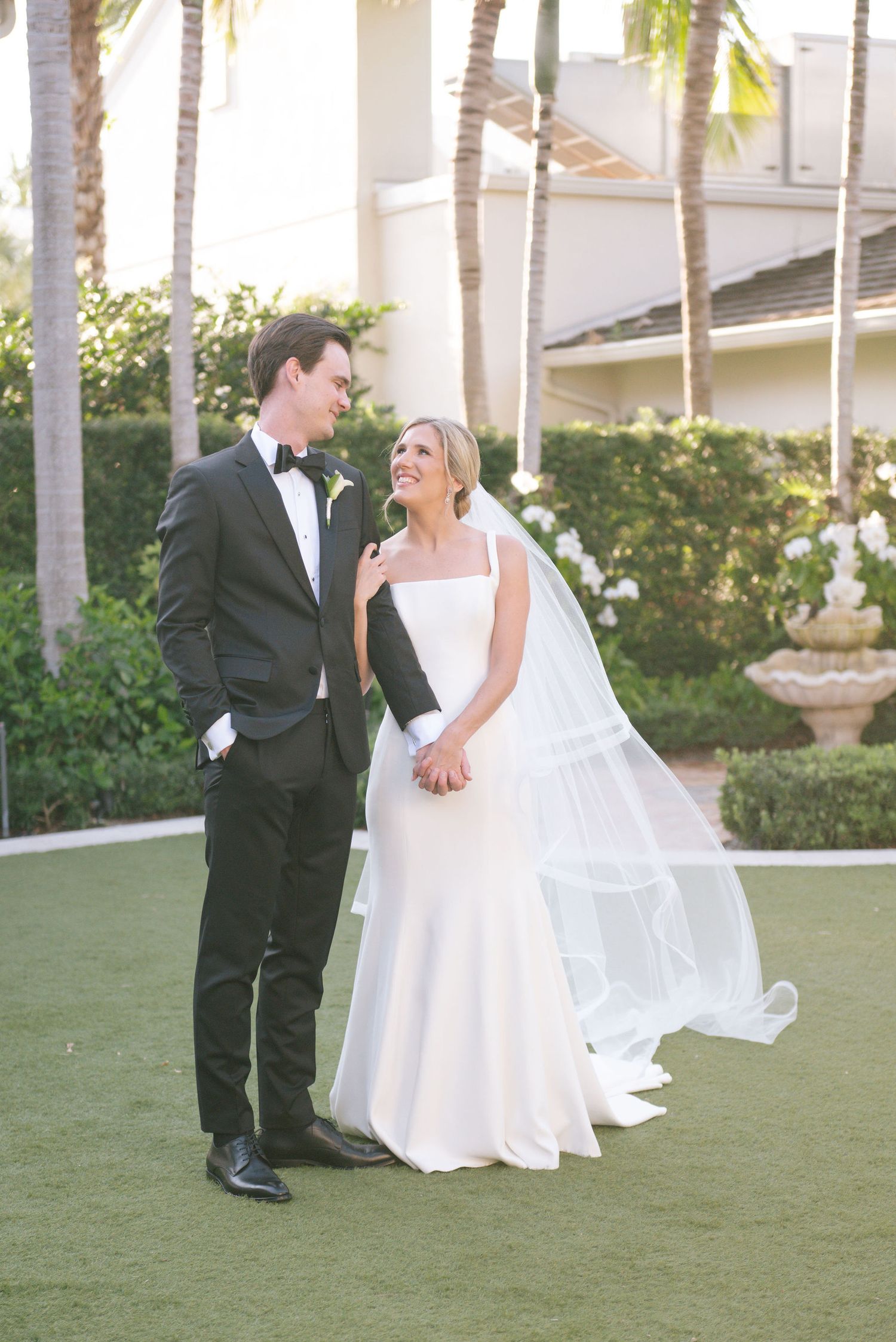A couple shares a romantic moment on a grassy lawn with palm trees and elegant architecture in the background.