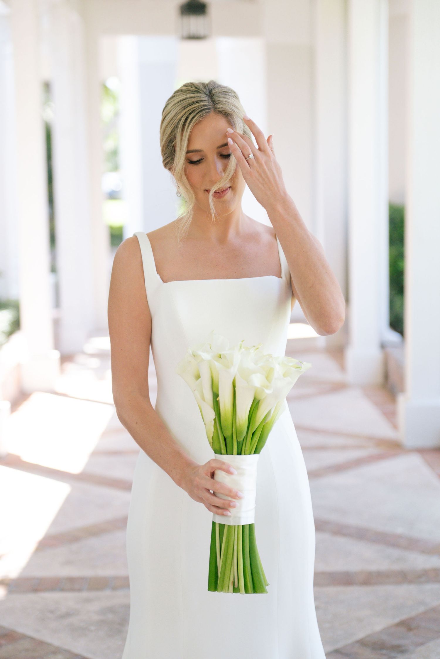 Bride in white dress holding a classic calla lily bouquet in a sunlit colonnade.