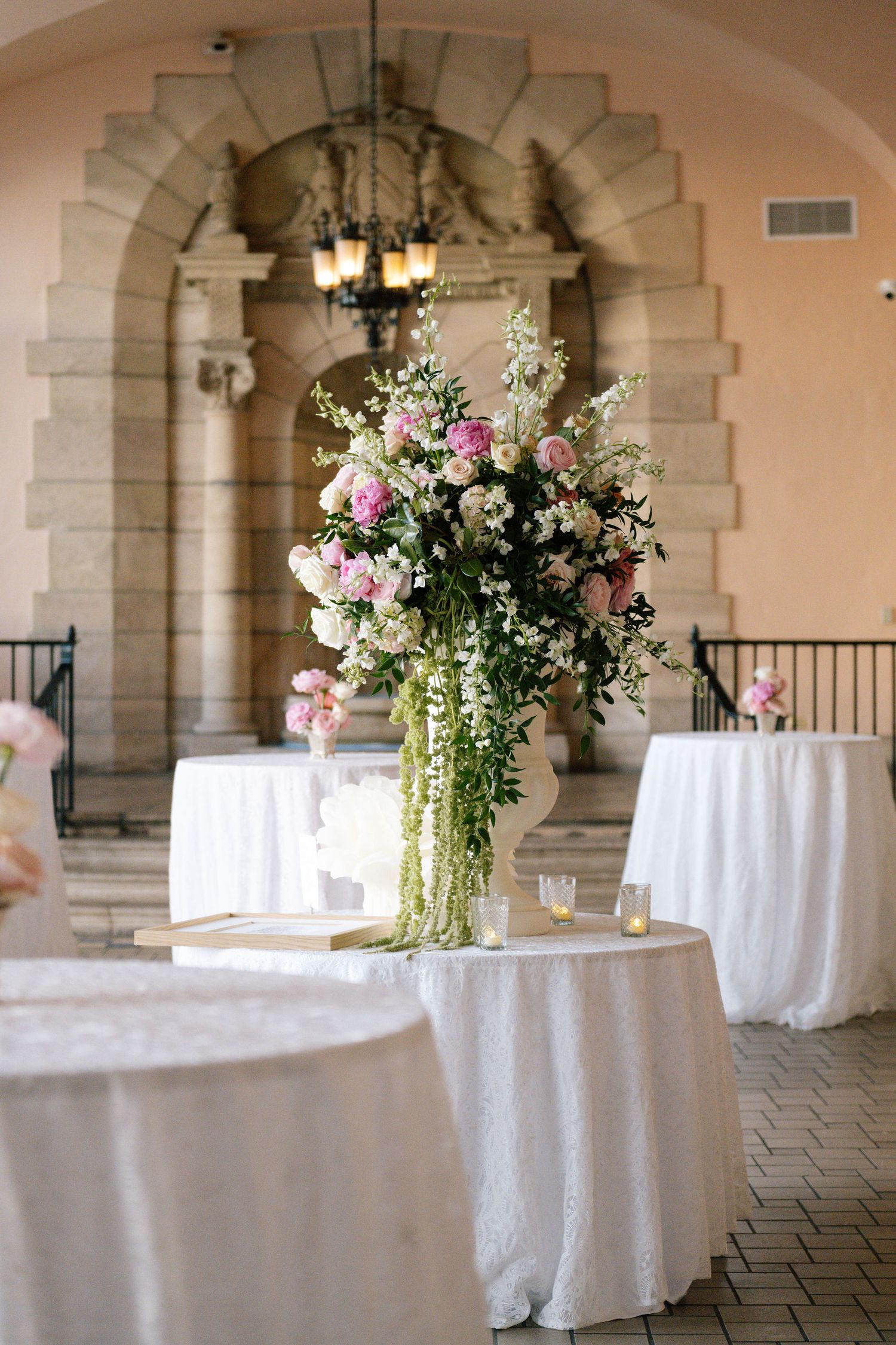 Round reception tables with white linens and pink floral centerpieces in an arched venue.