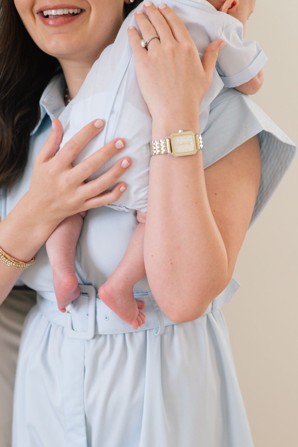 Woman in light blue dress holding baby while wearing gold jewelry and watch.