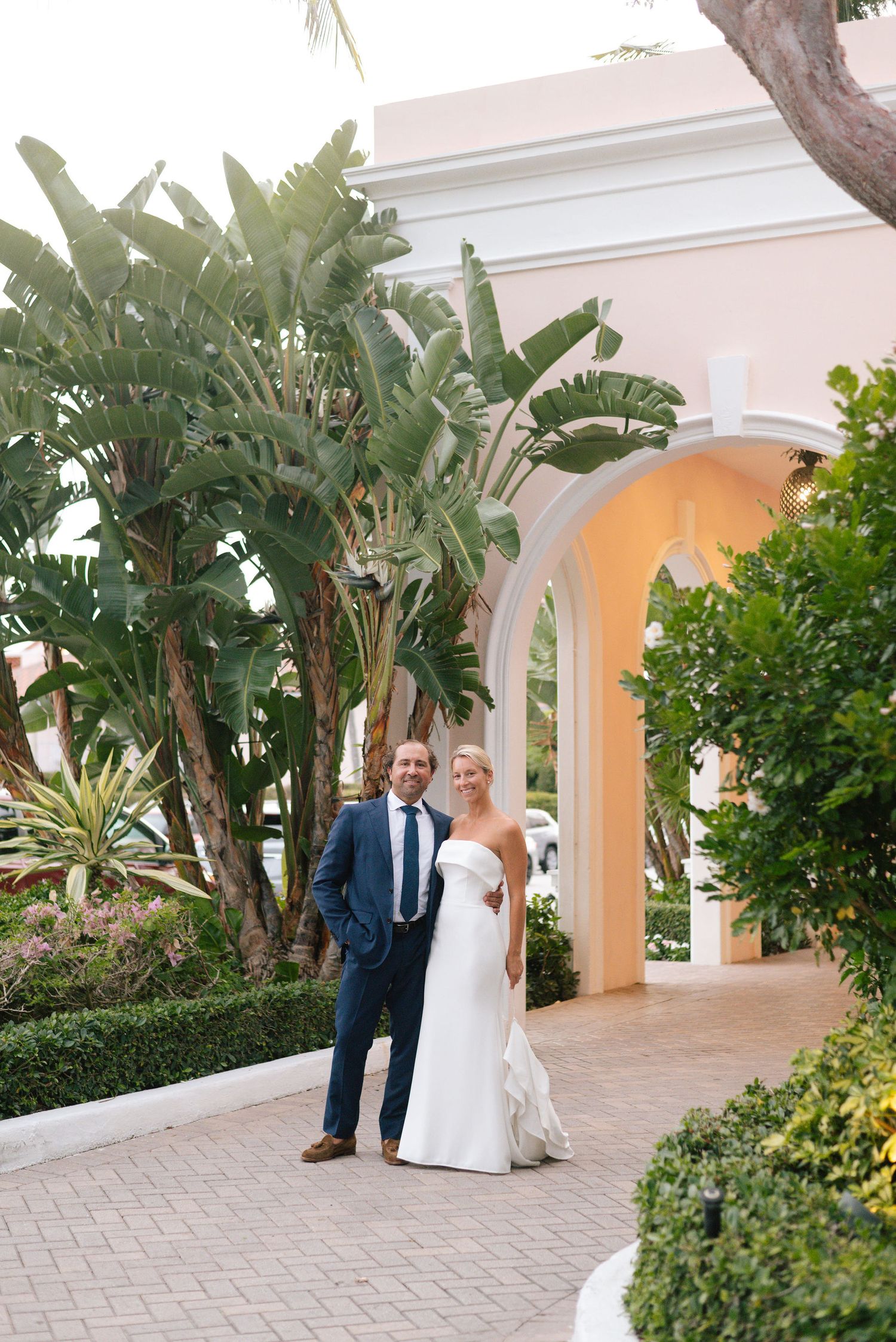Couple walking along a pink-walled garden pathway with tropical landscaping and arched columns.