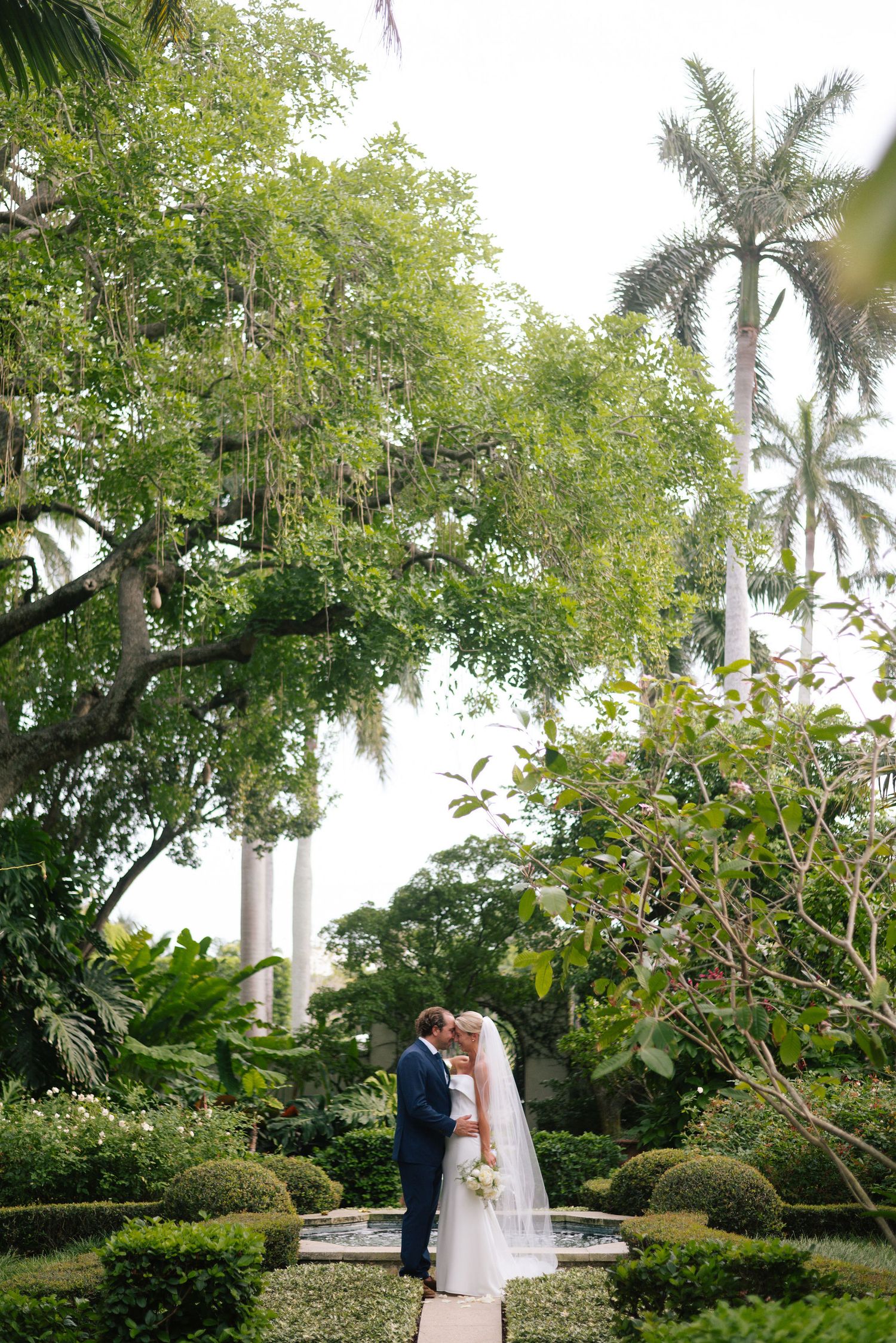 Wedding couple embracing in an elegant garden setting surrounded by palm trees and greenery.