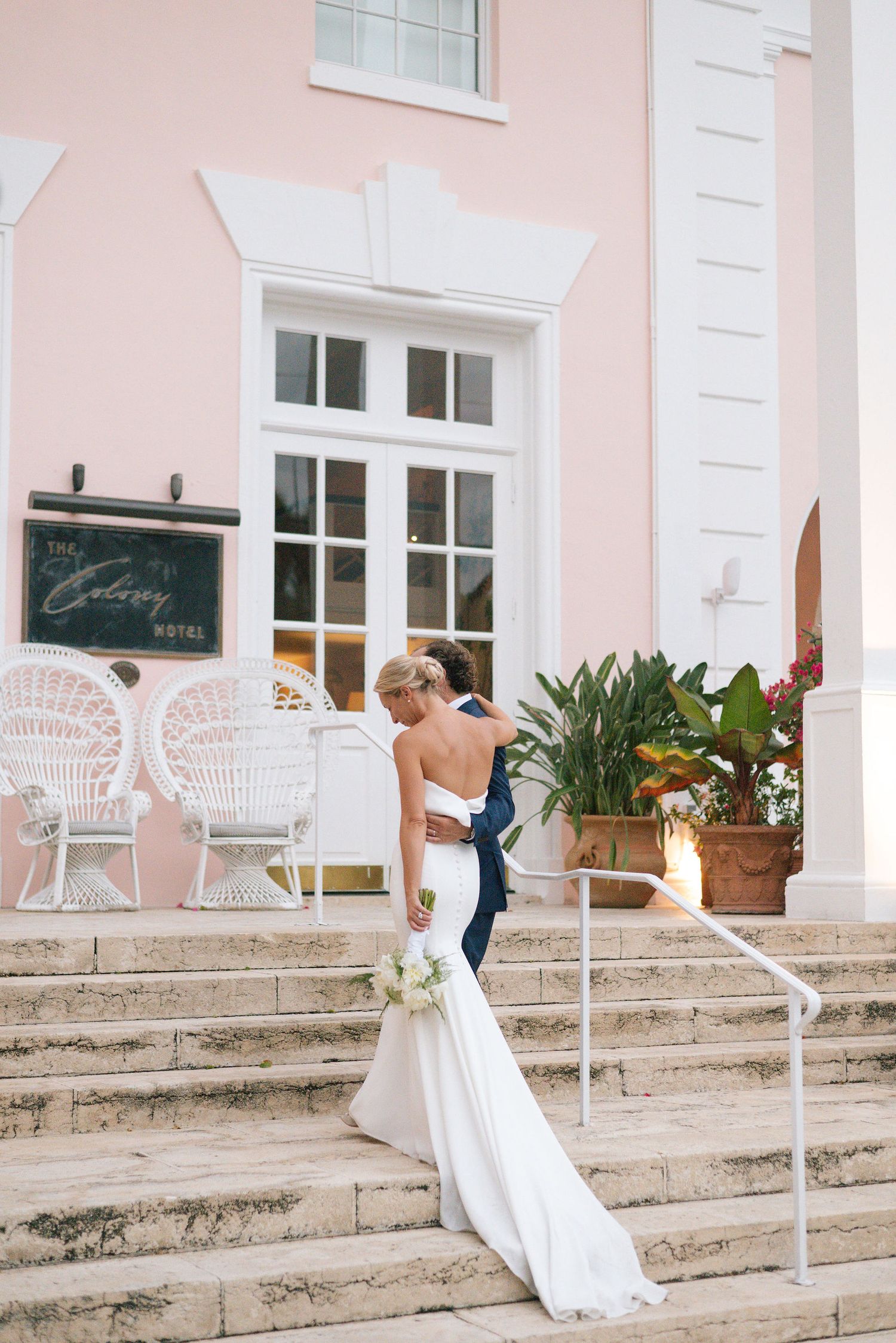 Wedding couple on stone steps with white wicker chairs and tropical plants at luxury hotel entrance.