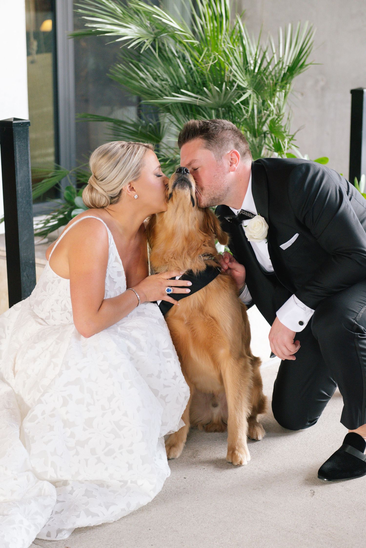 Bride and groom kissing their golden retriever dog during wedding day photos outdoors.