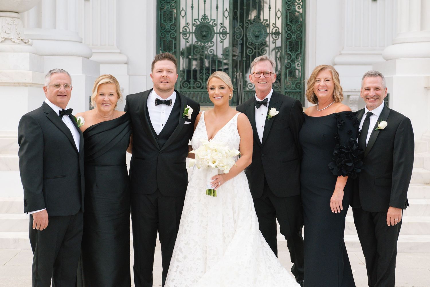 Wedding party in black formal attire poses with bride in white gown holding bouquet on elegant venue steps.
