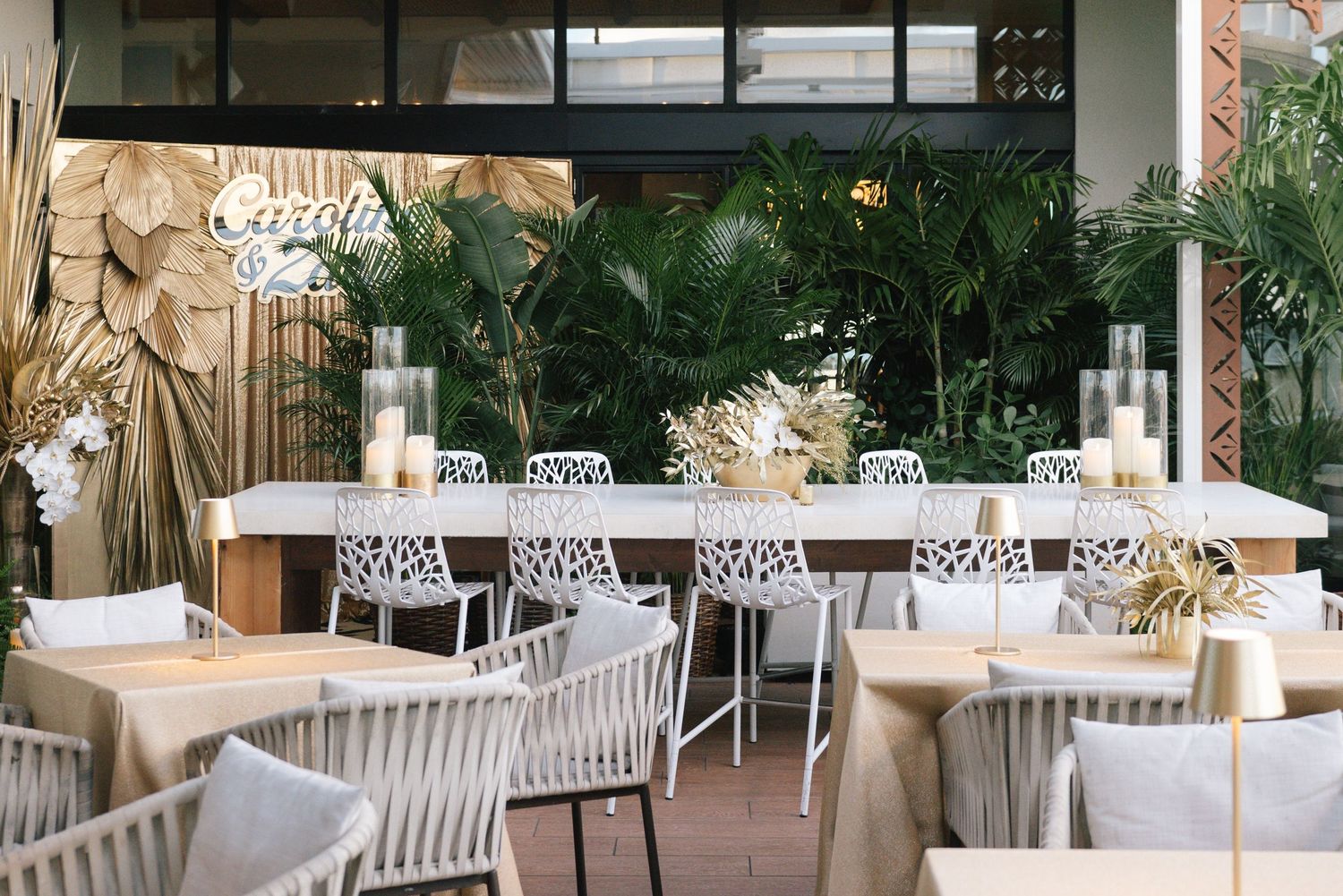Modern restaurant interior with white chairs, long dining tables and tropical palm decor in an elegant setting.
