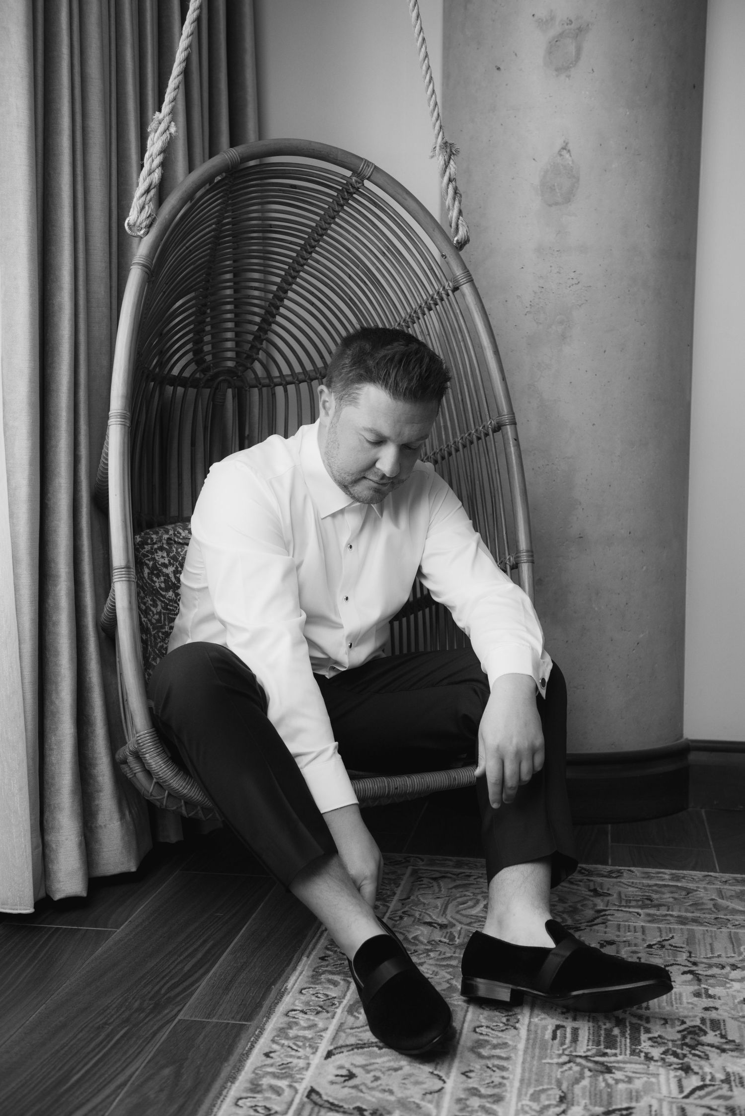 Man in white dress shirt and black pants putting on loafer shoes while seated in a hanging rattan egg chair.