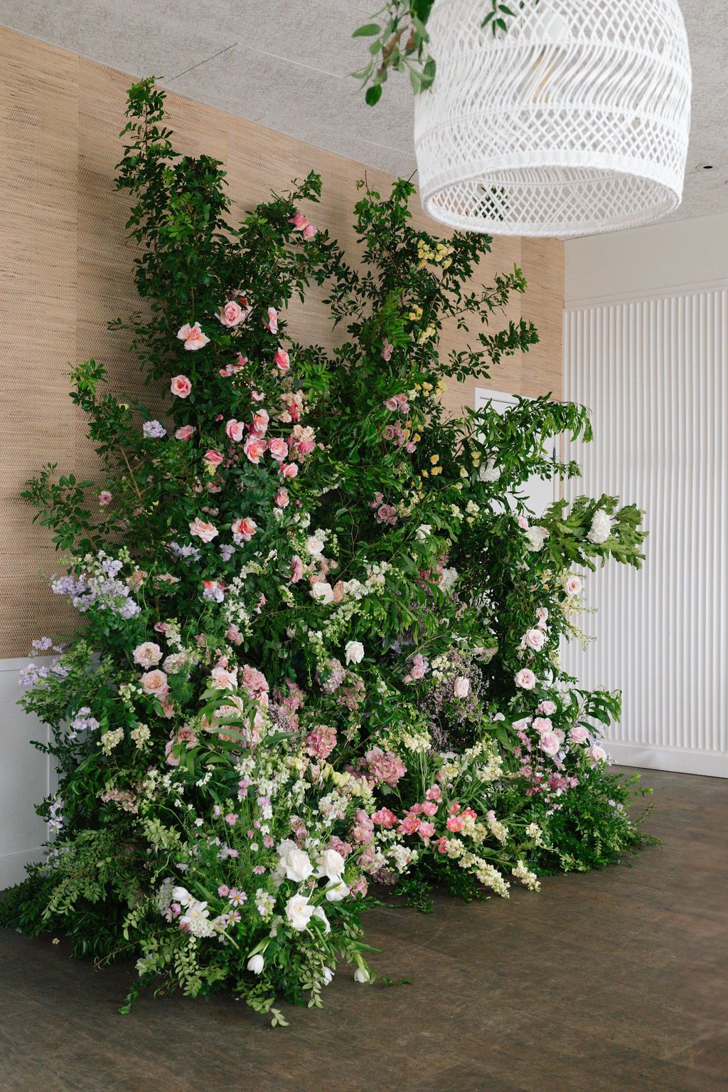 Lush floral installation with pink roses and greenery against a beige wall with white pendant light fixture.