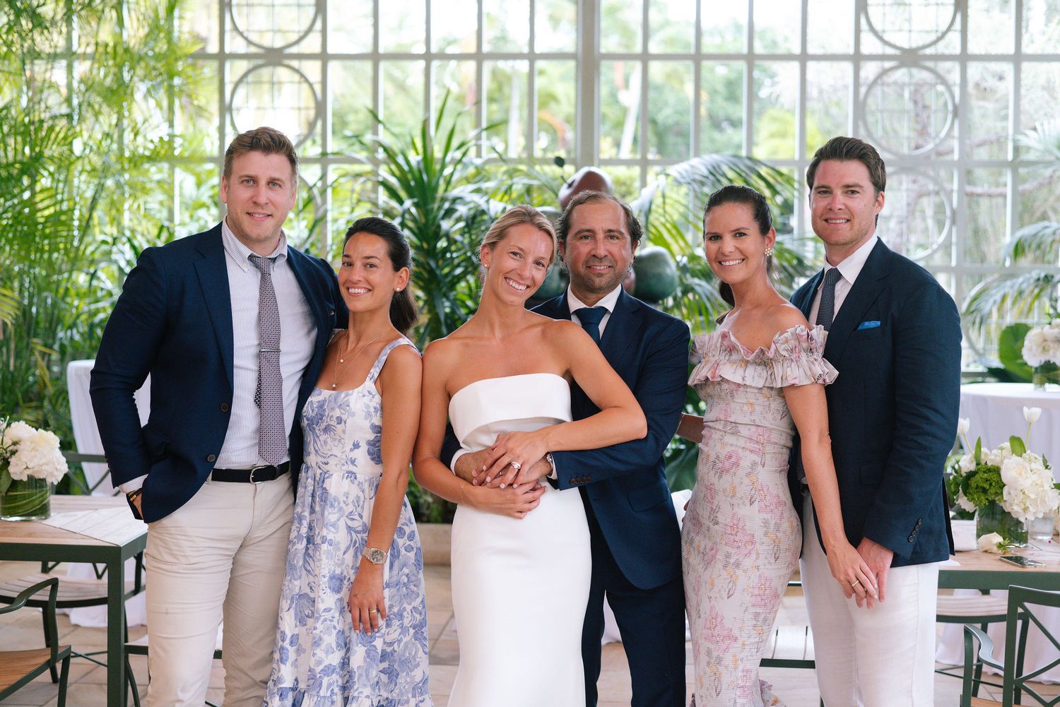 Wedding party in formal attire poses together in an elegant indoor venue with tropical plants.
