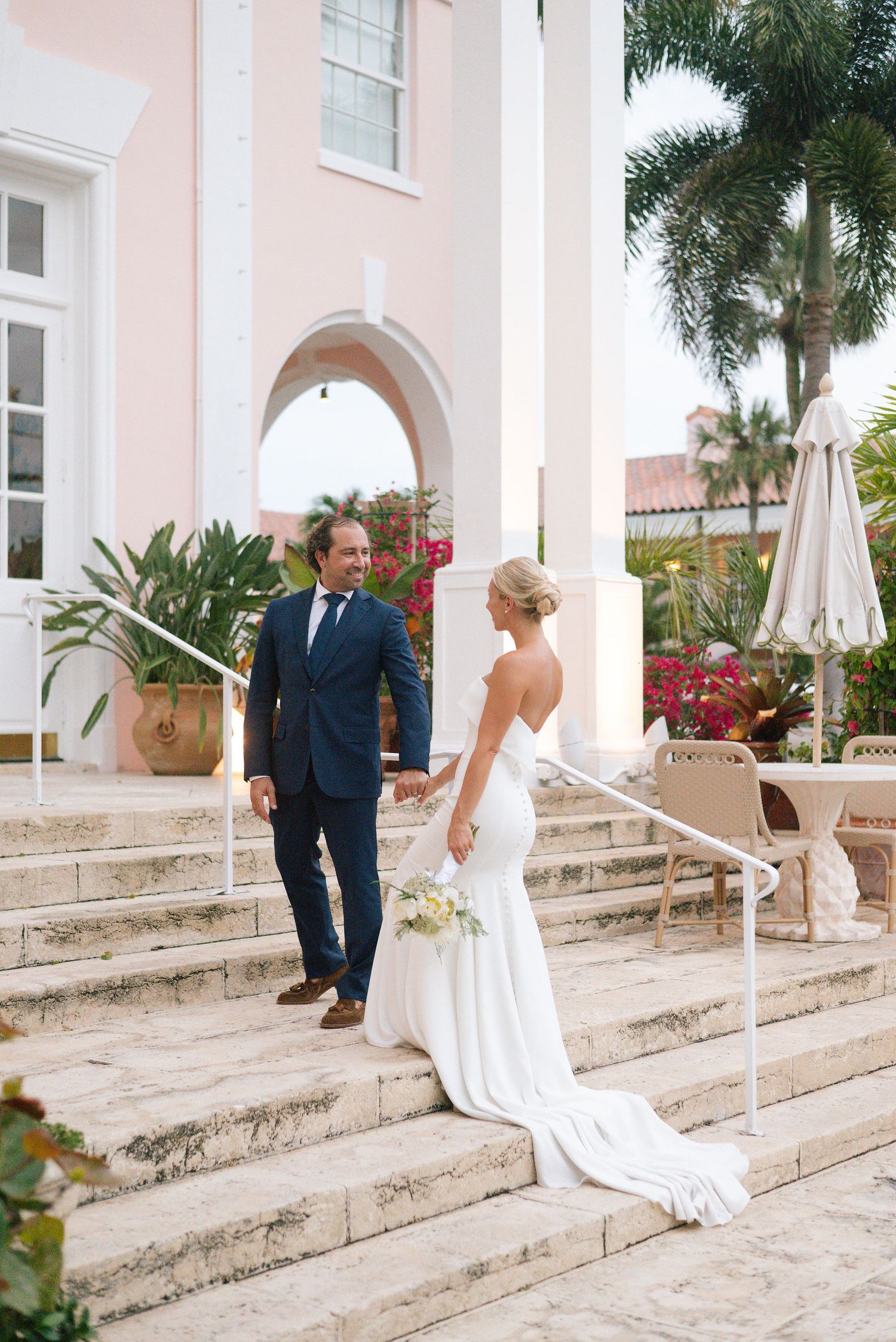 Couple shares a first look moment on stone steps of a pink colonial-style building.