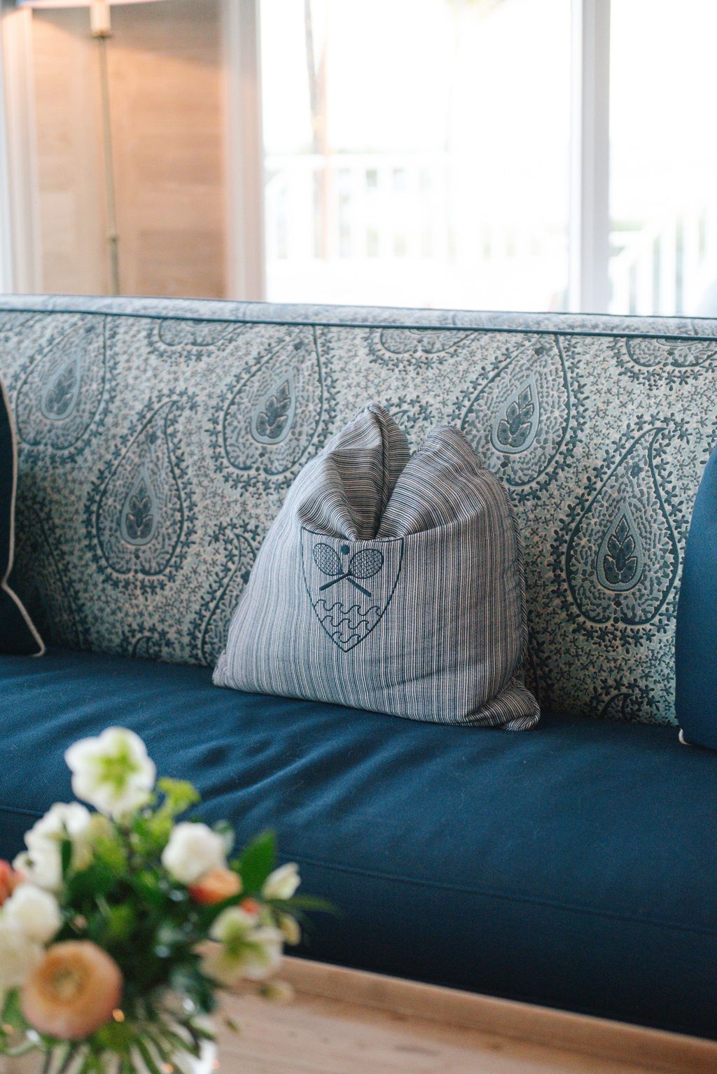 Blue sofa with paisley pattern cushions and decorative gray pillow next to fresh flowers.