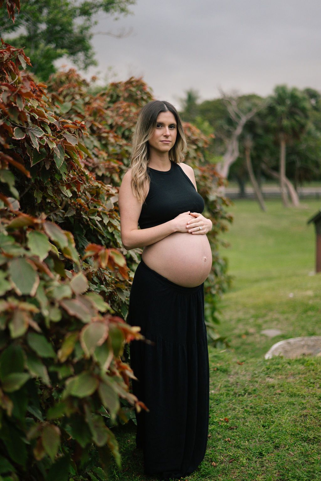 Maternity photos showcase woman in black dress standing among autumn foliage.