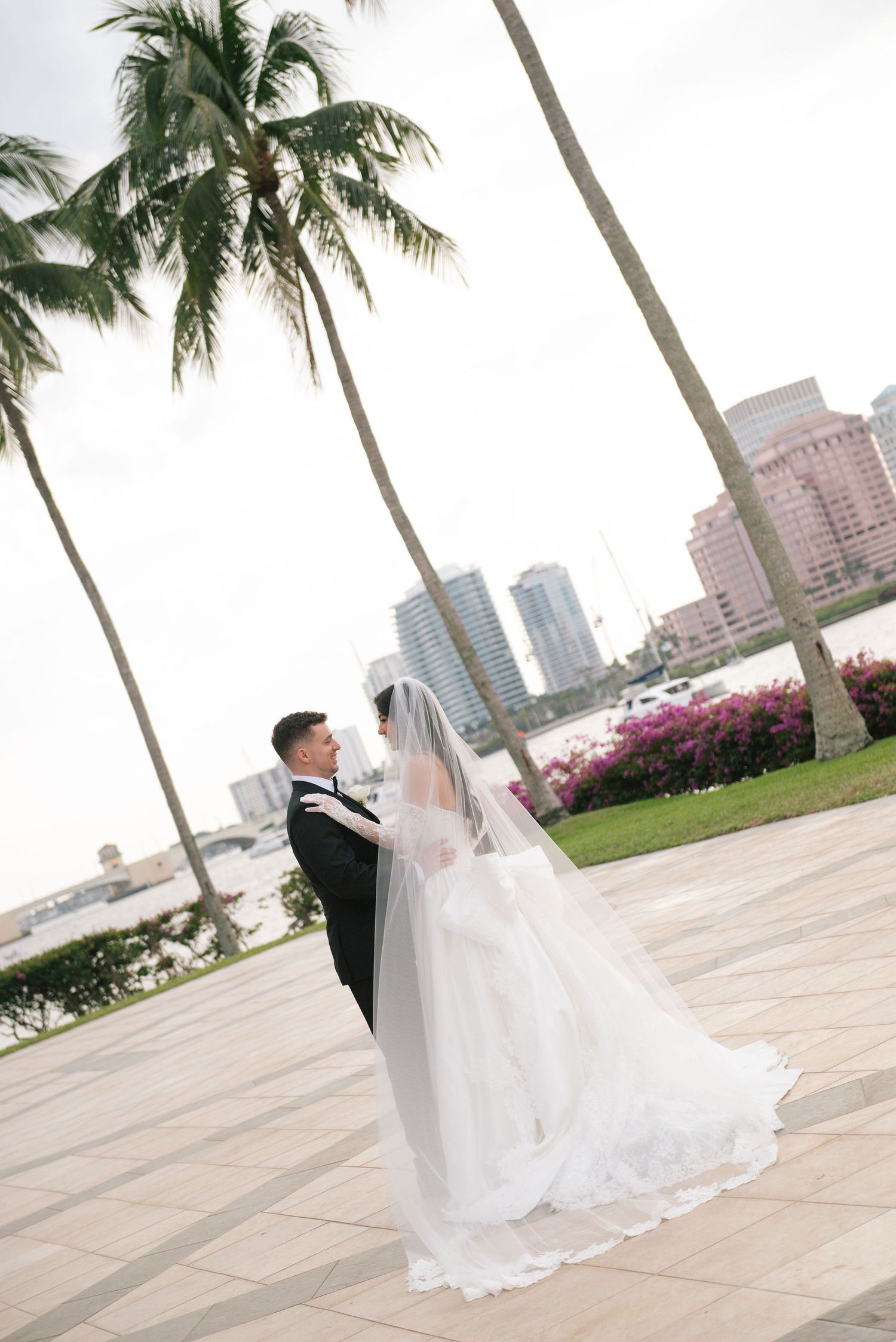 A wedding couple poses elegantly against a backdrop of palm trees and city buildings in an outdoor setting.