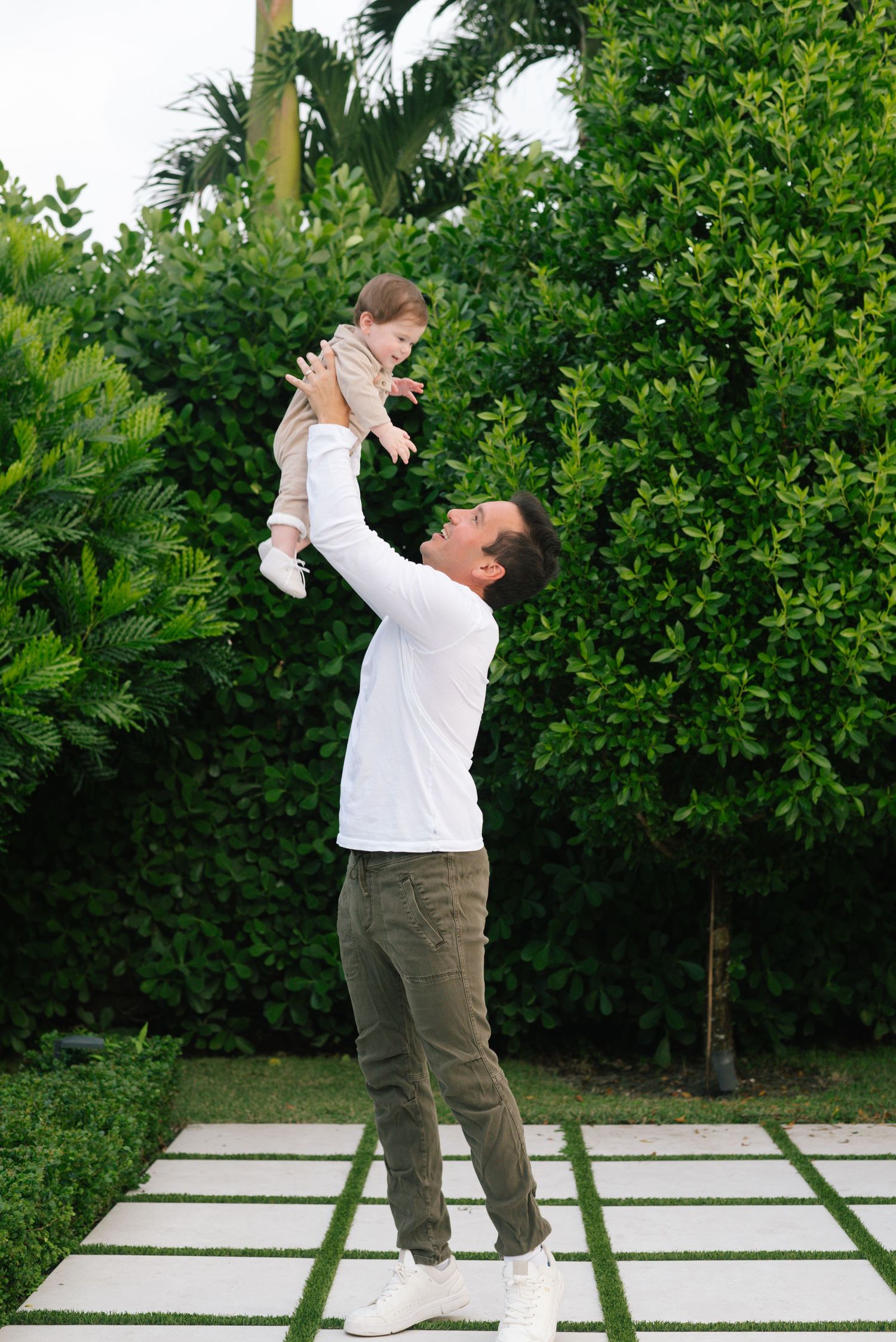 A parent in a white shirt lifts a small baby high into the air while standing on concrete pavers in a lush garden setting.