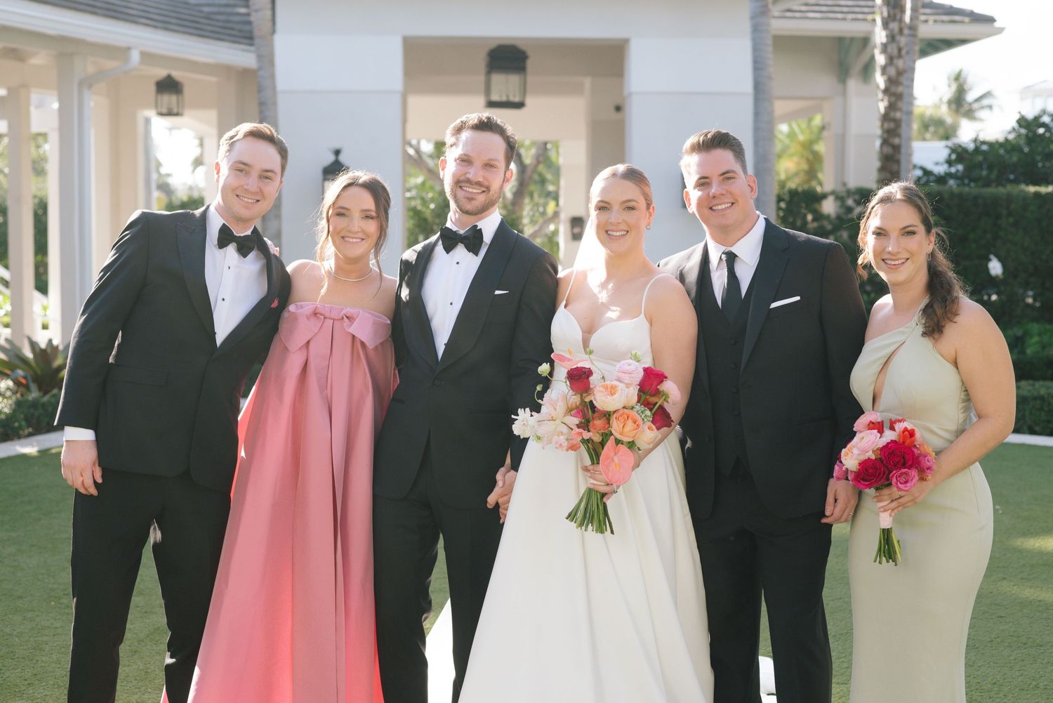 Wedding party in pink bridesmaid dresses and black tuxedos posing for formal photos outdoors.