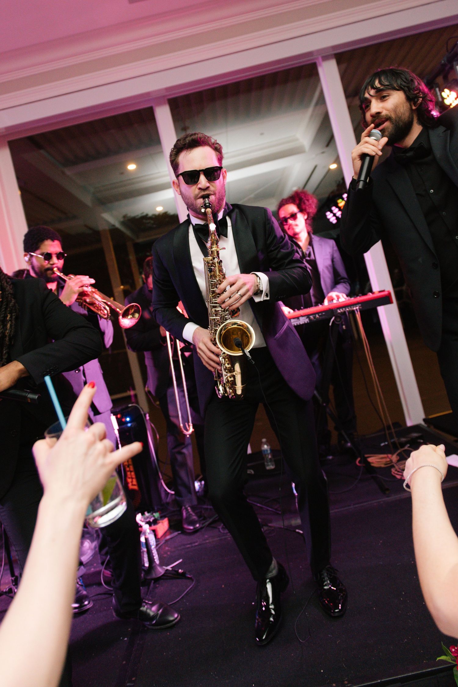 A band performs at a wedding reception with musicians in black suits playing instruments under pink lighting.
