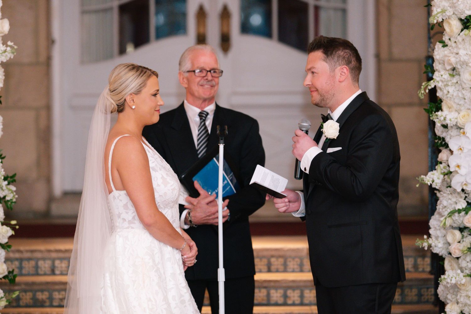 Bride and groom exchange vows at elegant wedding ceremony with officiant and white floral arch backdrop.