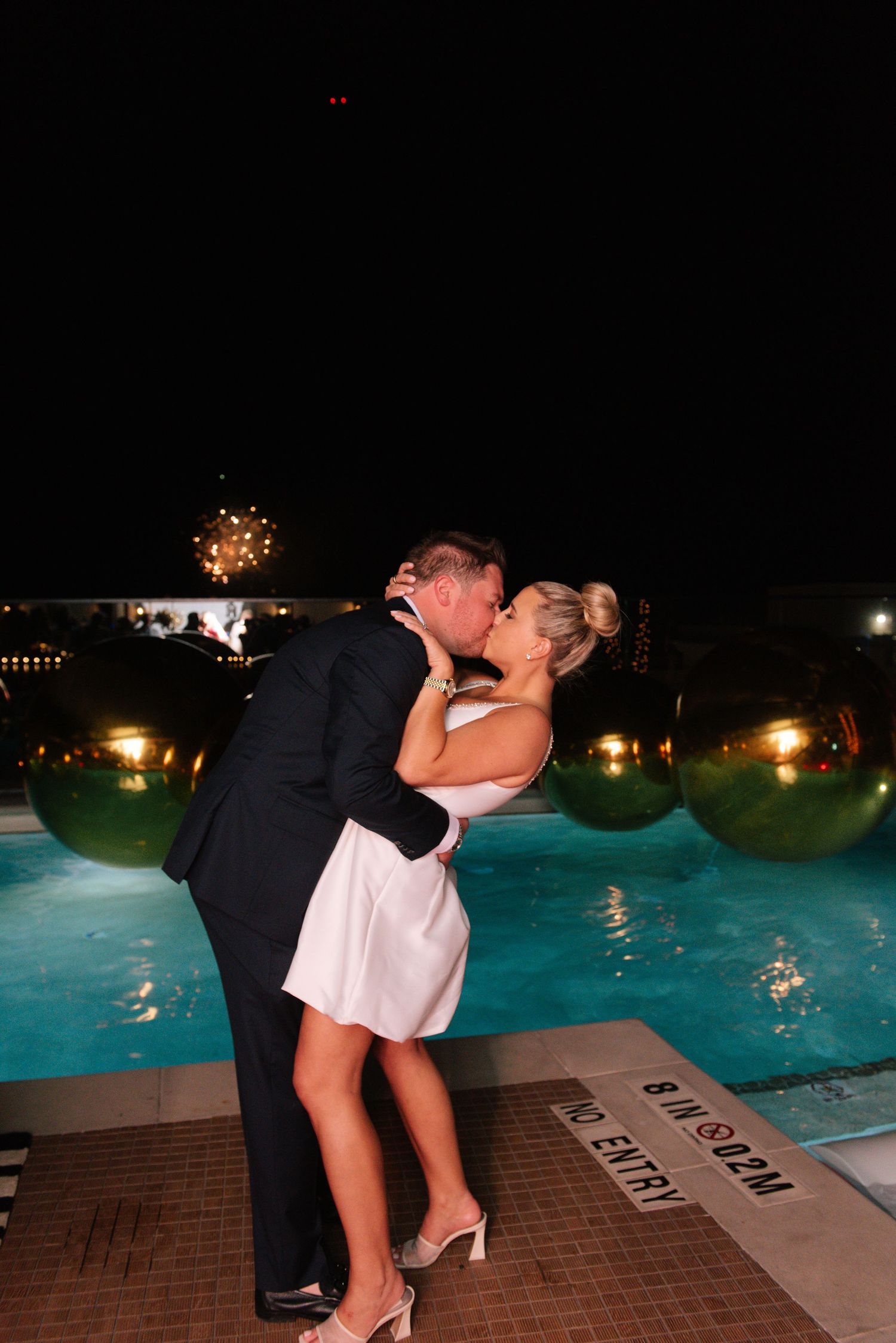 A romantic couple sharing a kiss by an illuminated swimming pool at night.