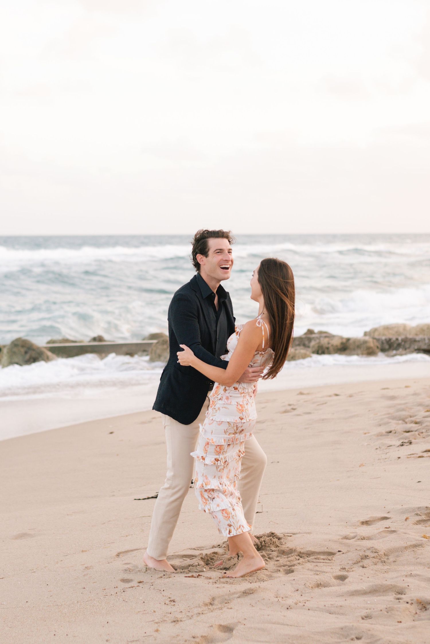 A couple shares a romantic moment on a sandy beach with crashing waves and rocks in the background.