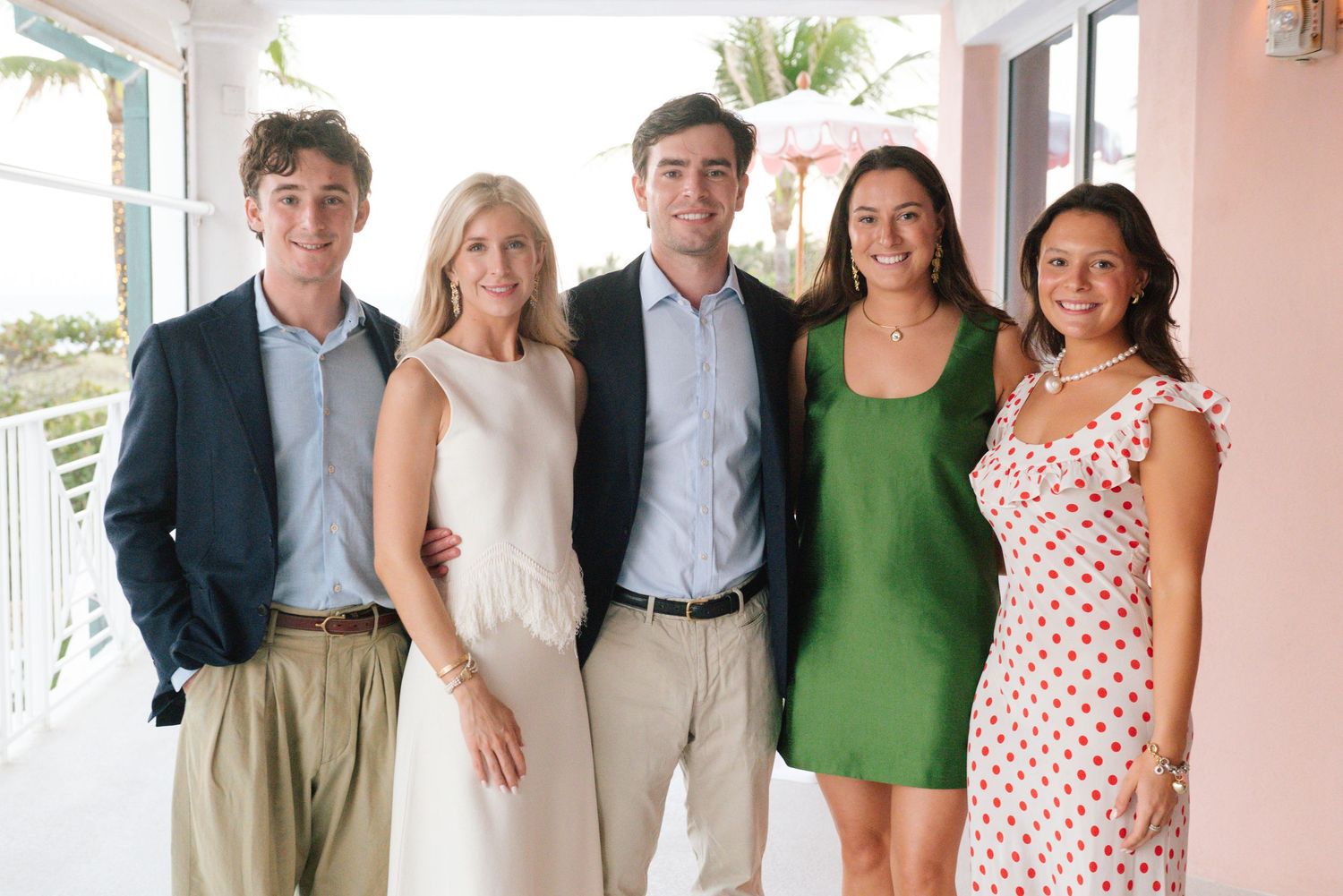 A group of five people stand together smiling on a covered porch wearing summer attire in casual formal wear.