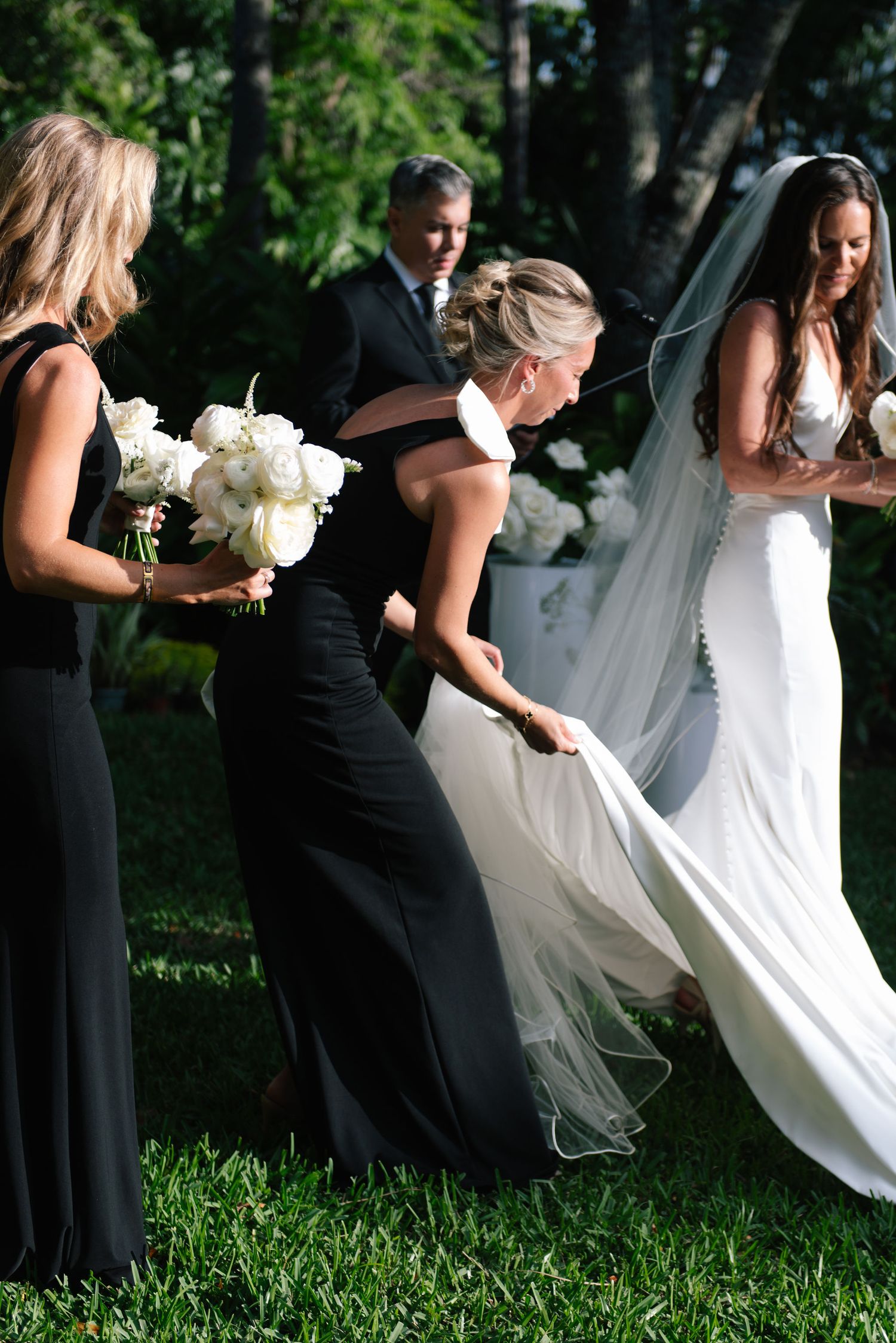 Wedding party members in black dresses help adjust a white wedding gown on grass.