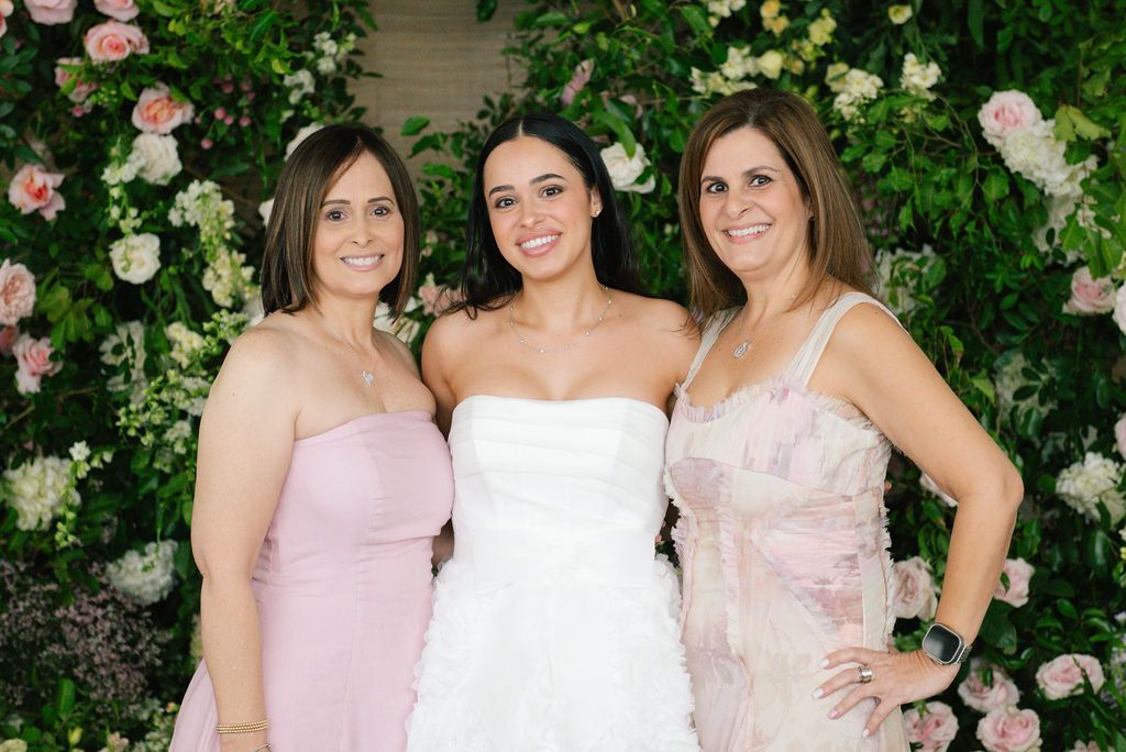 Wedding party group photo with soft pink dresses against a floral wall backdrop.