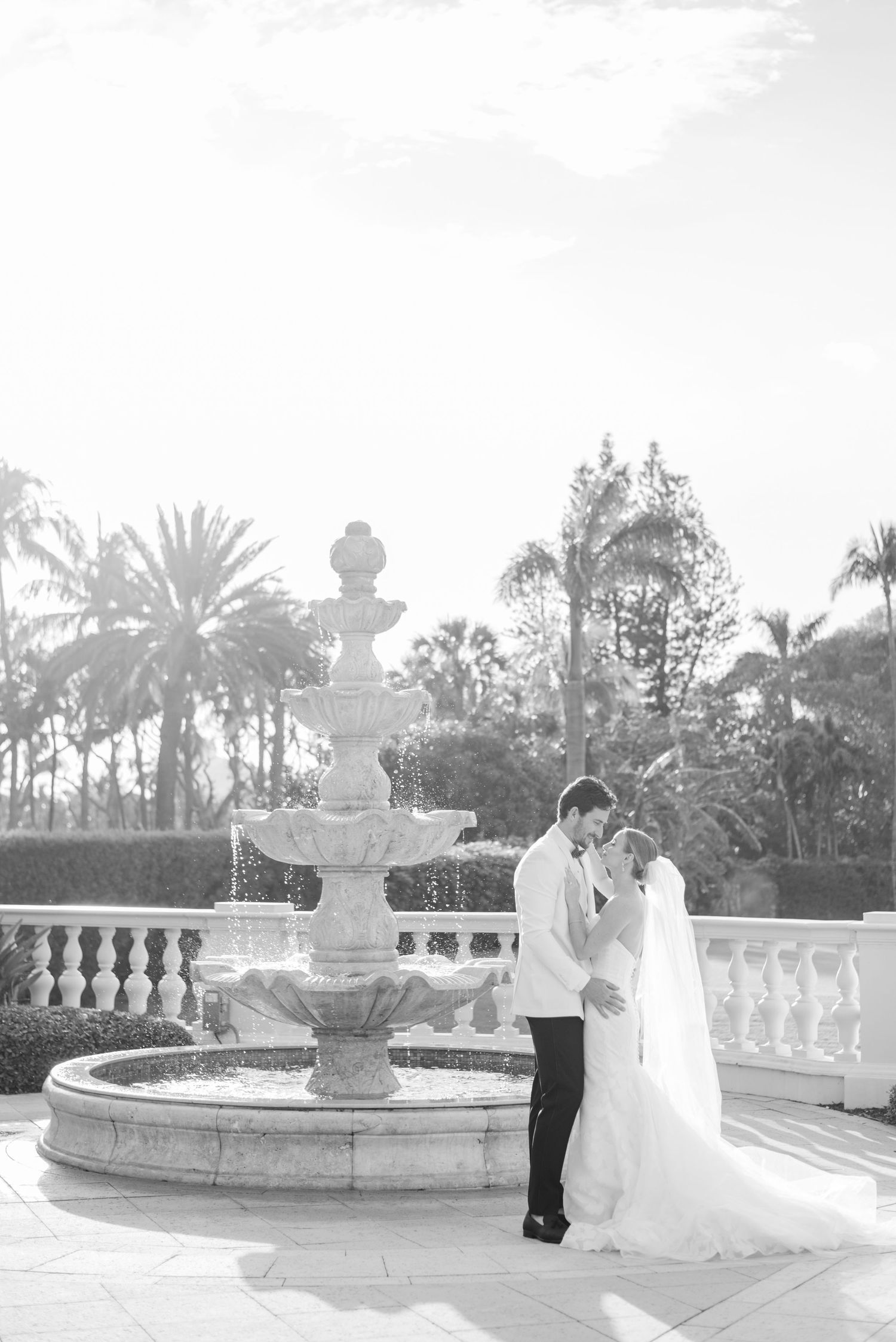 Newlyweds share tender moments by an ornate fountain surrounded by palm trees.