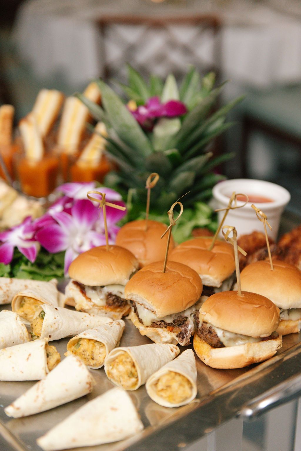 Catering display of mini sandwiches and sliders garnished with tropical flowers and greenery.