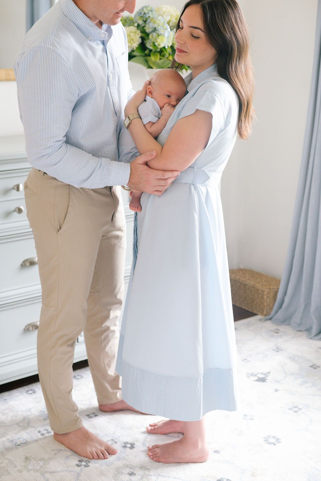 Parents standing barefoot on plush carpet share a tender moment with their newborn baby.
