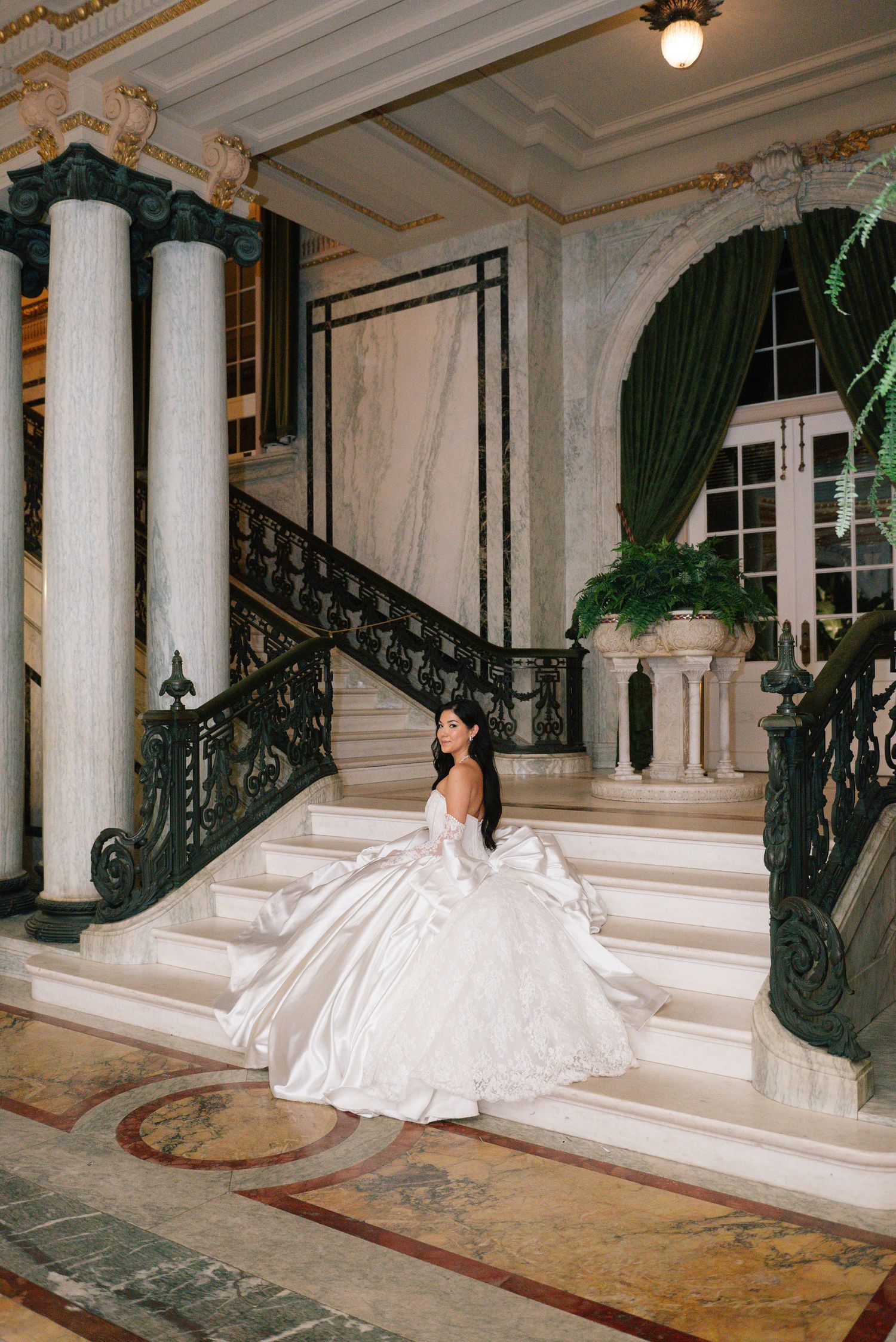 A bride in a white gown sits gracefully on marble steps in an ornate interior.
