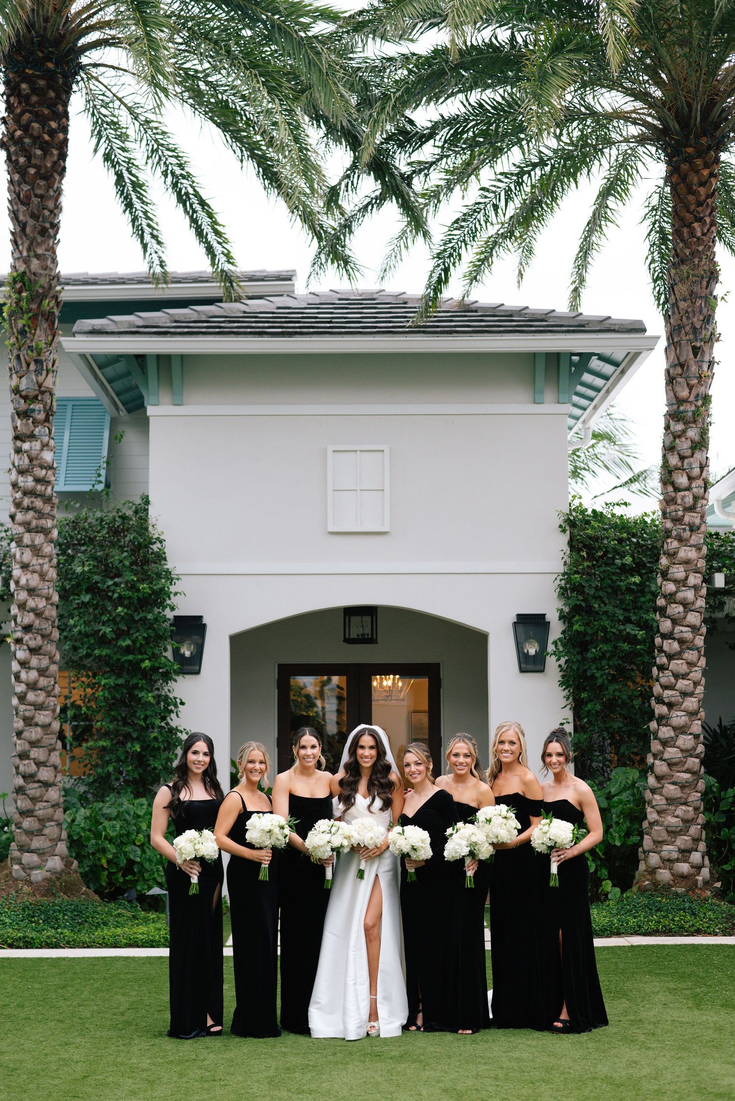 Wedding party in formal black and white attire lined up for photos in front of palm trees.