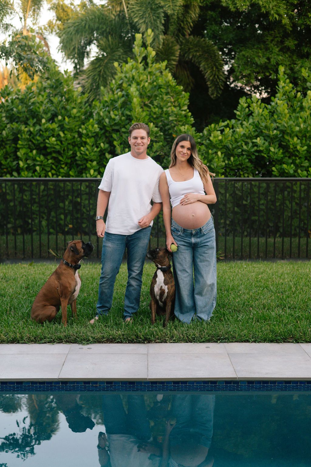 A couple stands by a pool with their brown dog in a backyard with green trees.