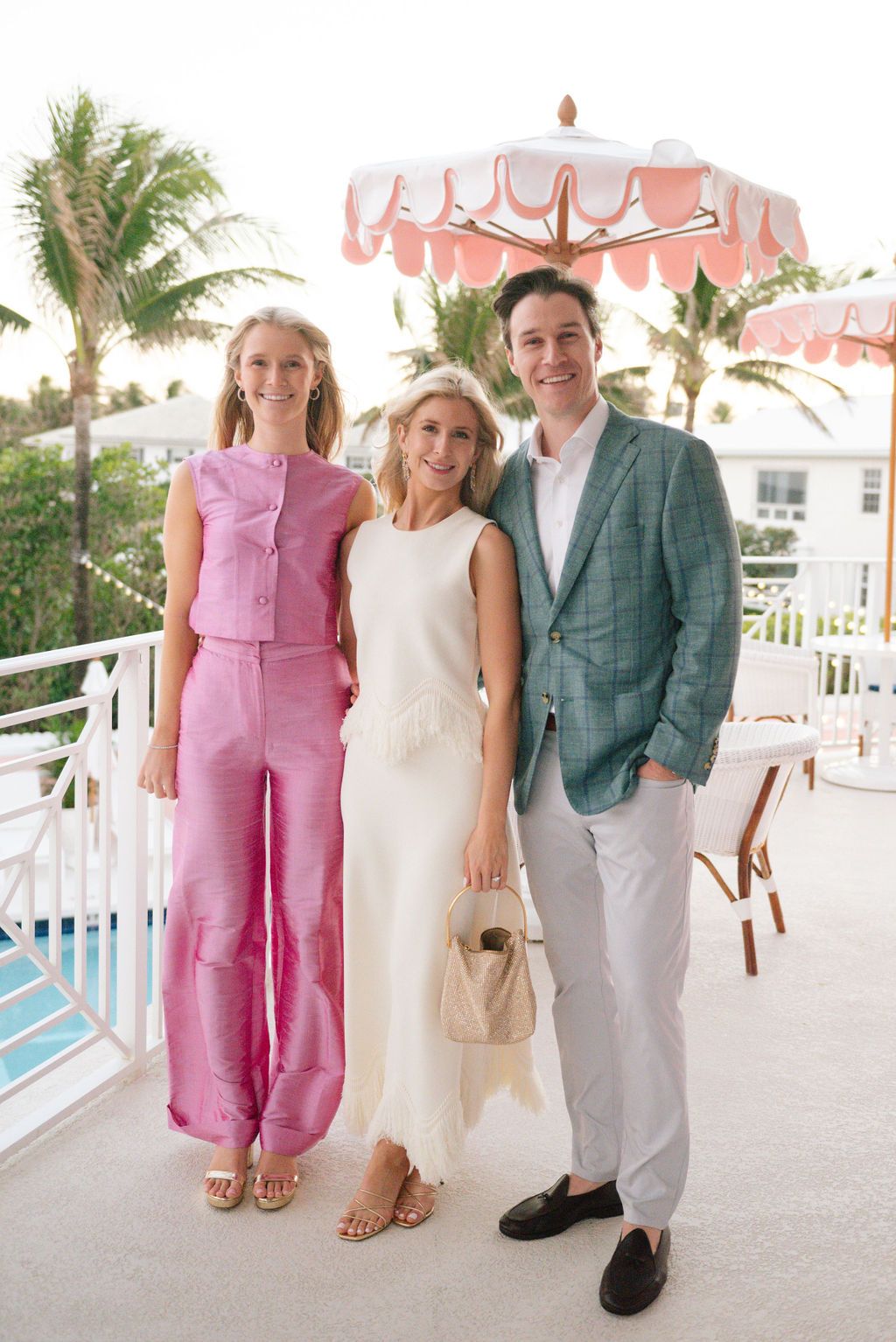Three people pose under pink umbrella on balcony wearing pink, white and green formal attire.
