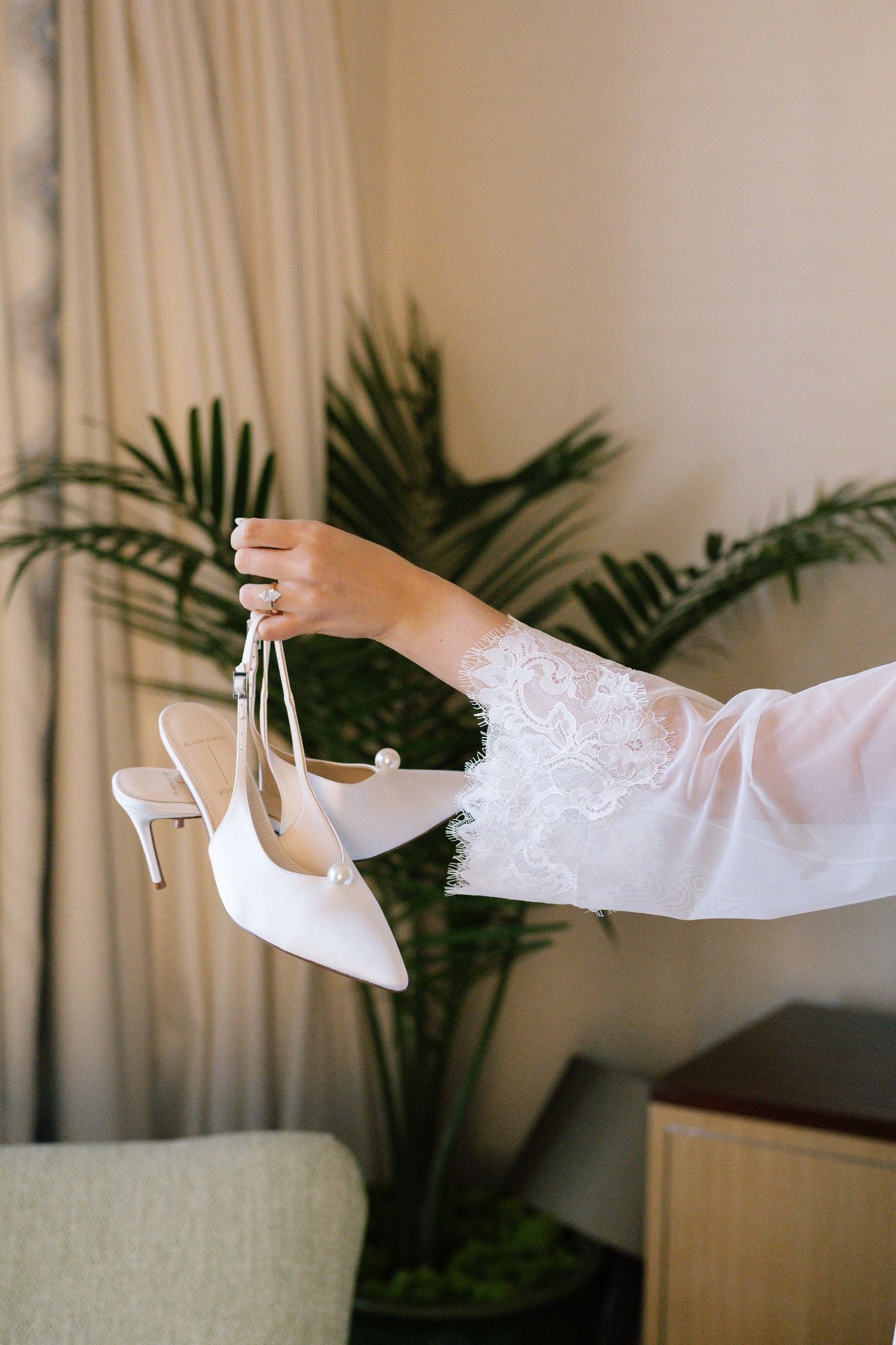 Hand holding elegant white bridal heels with palm plants in background.