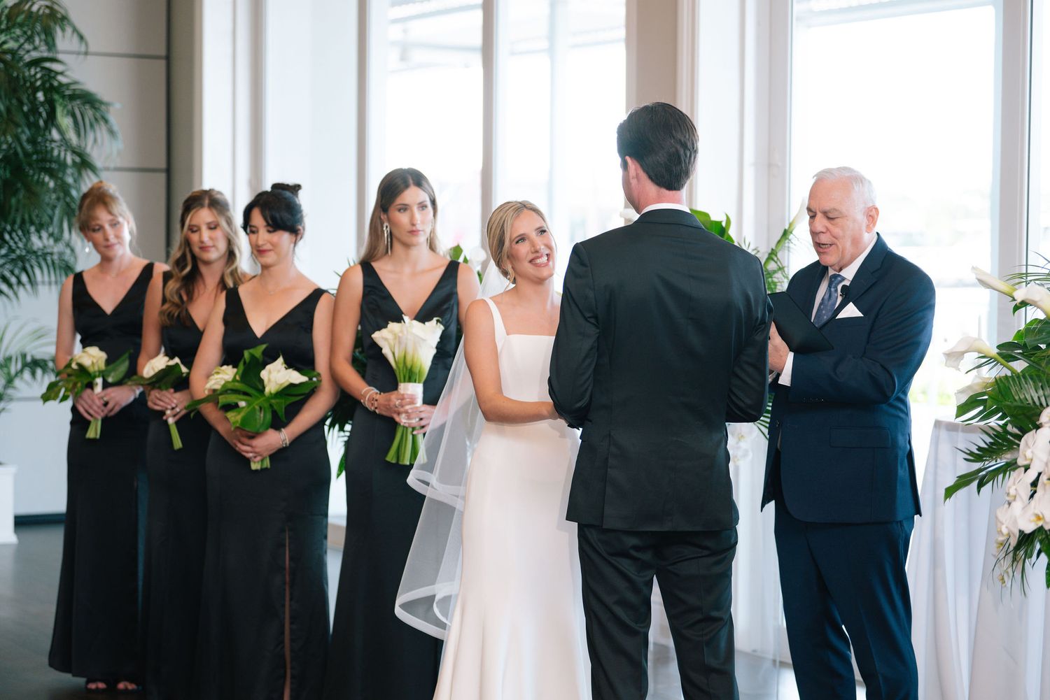Wedding party in black and white attire stands during indoor ceremony with tropical plants.