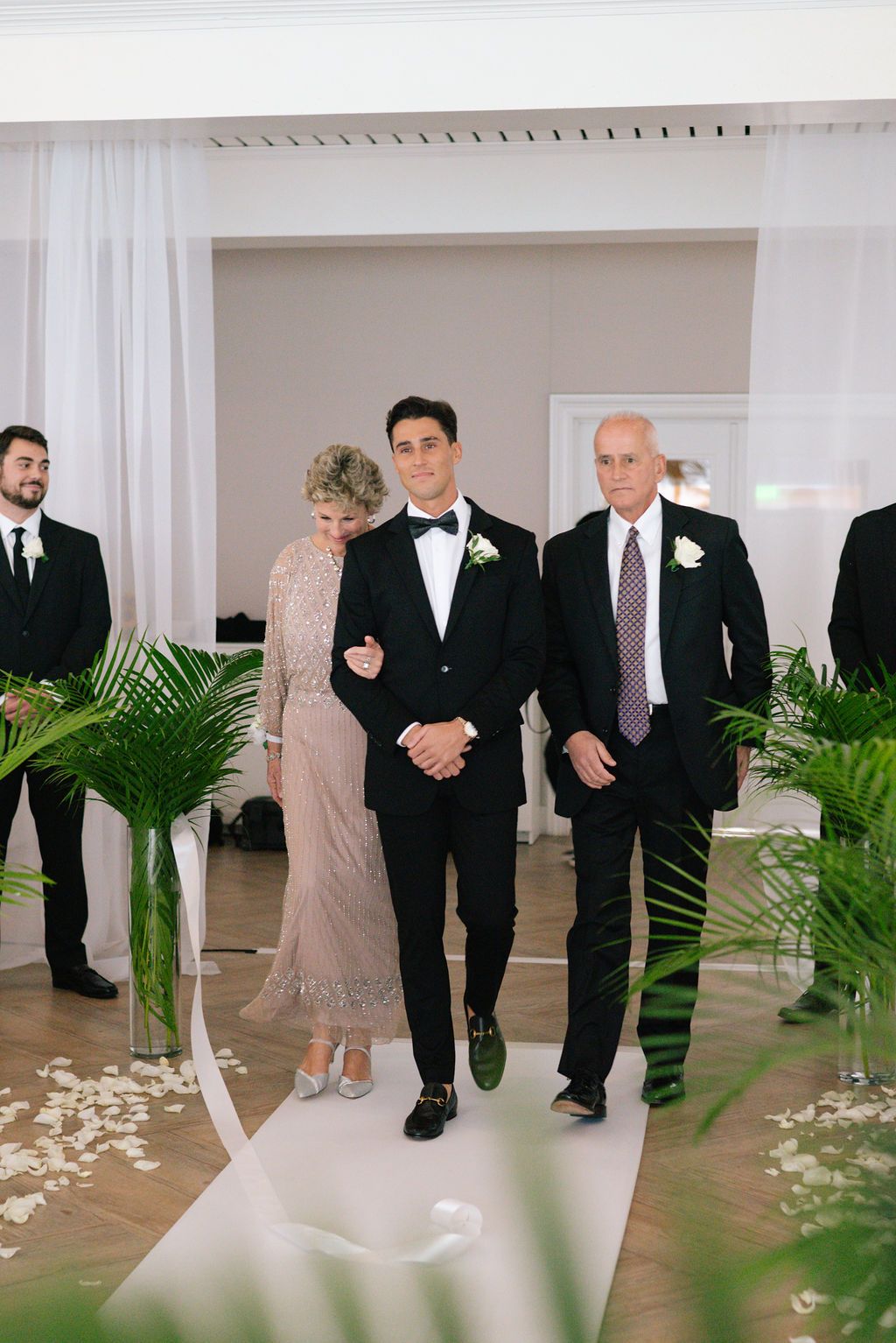 Wedding party members walking down the aisle decorated with palm plants and rose petals.