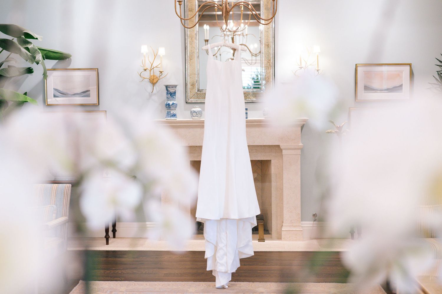 Elegant white wedding dress hanging in a bright room with crystal chandelier and soft lighting.