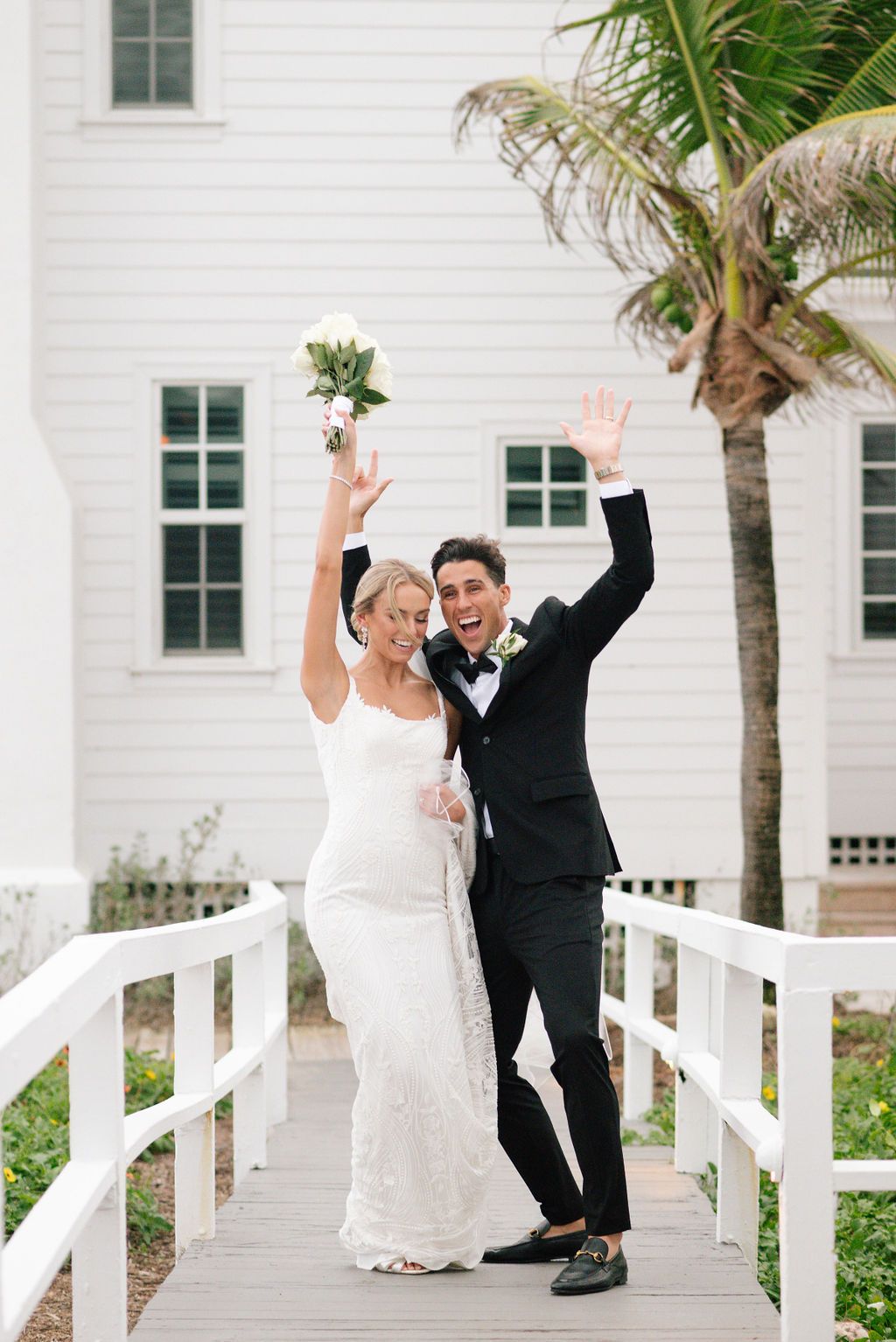 Wedding couple celebrates with raised arms on white boardwalk with palm trees.