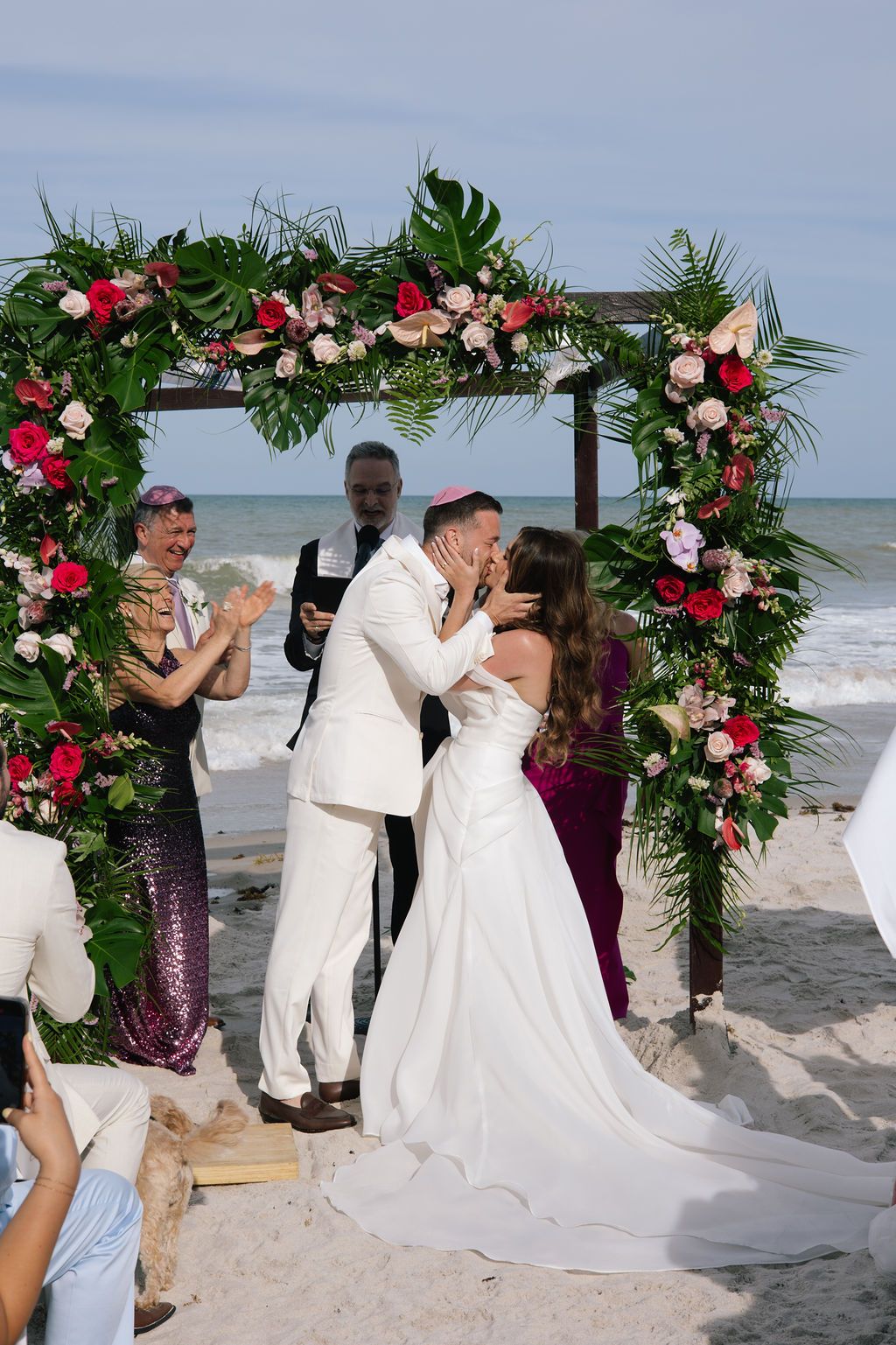 Wedding party celebrating under tropical floral arch on sandy beach with ocean views.