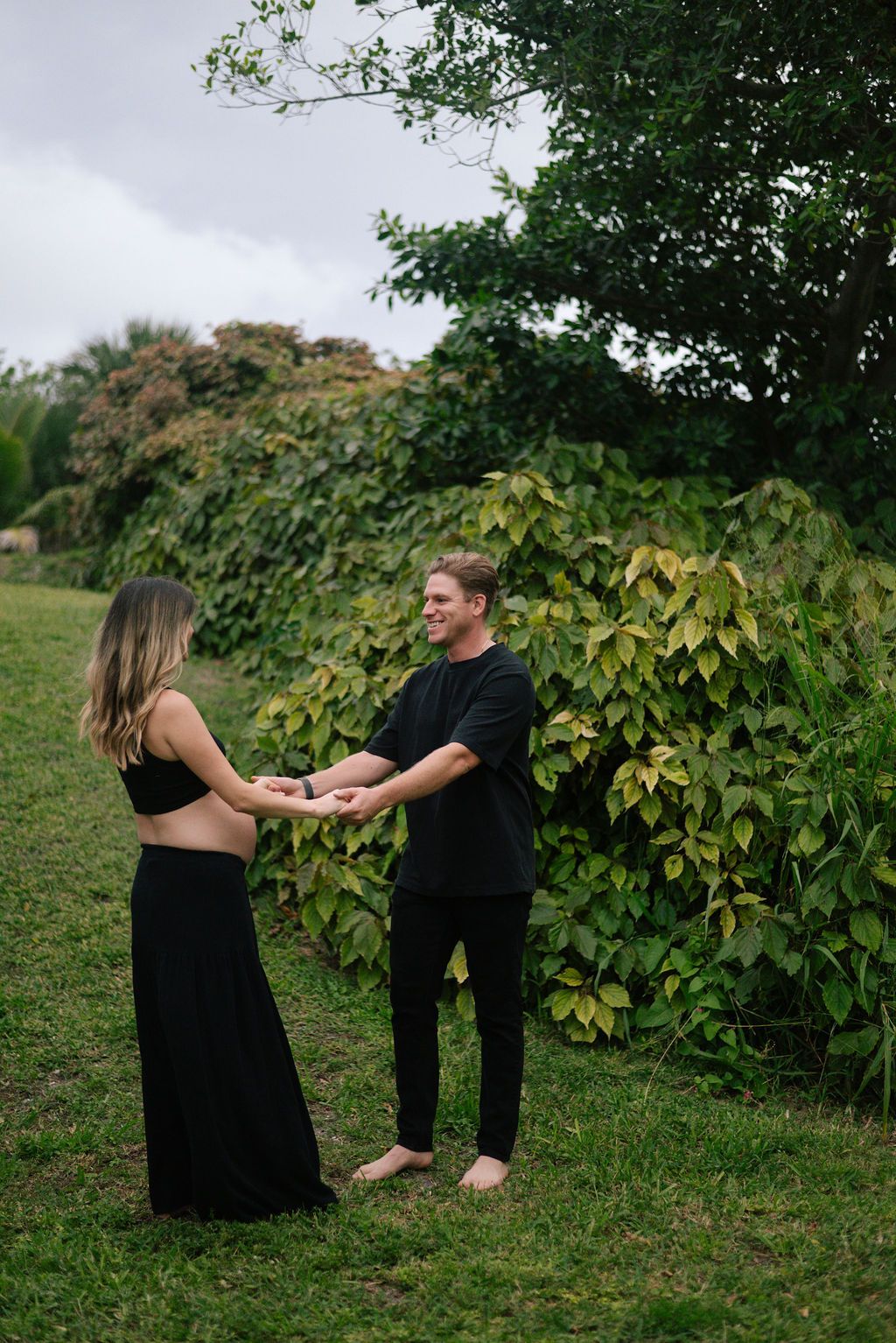 Couple holds hands and dances together on a grassy slope with leafy trees behind them.