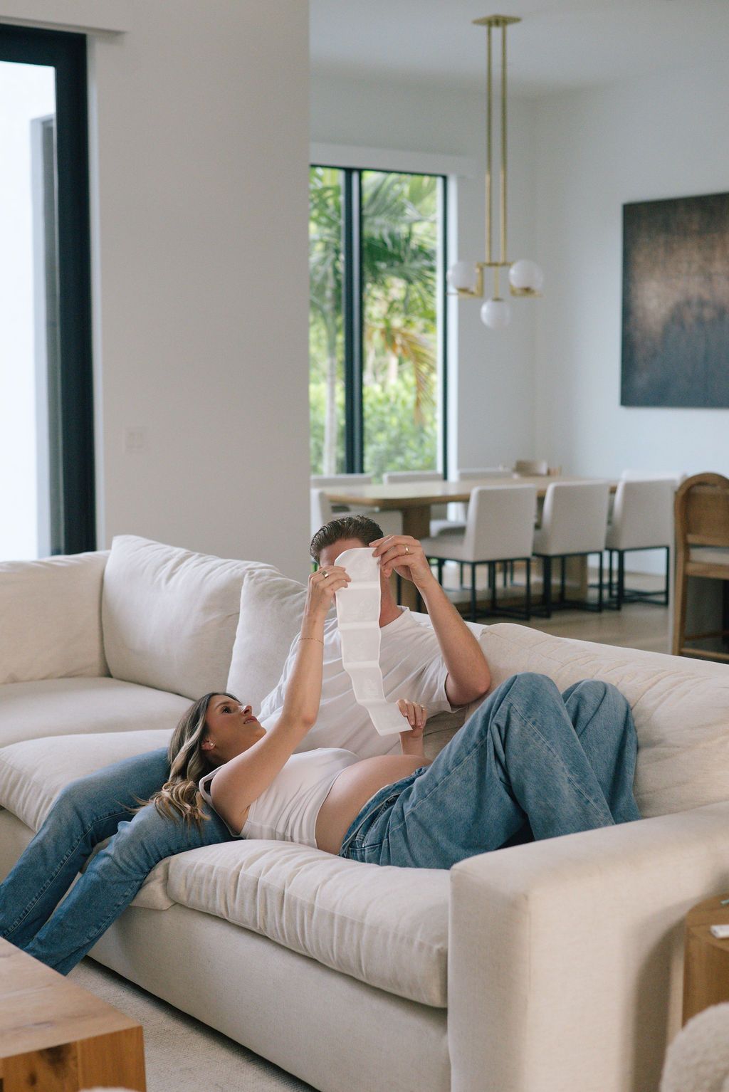 Person lying on white couch looking at ultrasound photos in bright modern living room.