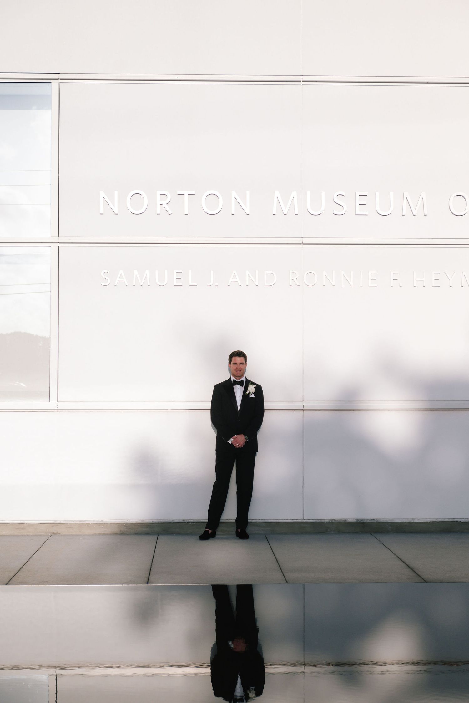 A groom in a tuxedo stands against the white wall of the Norton Museum with a mirrored reflection below.