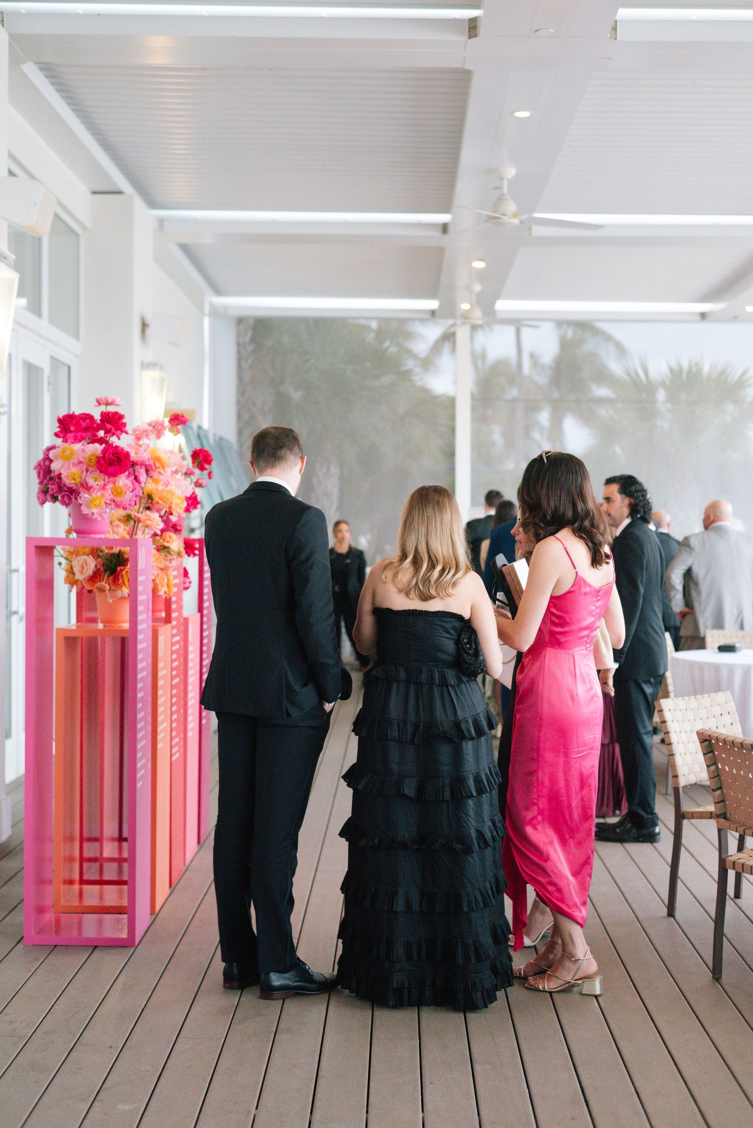 Wedding guests in formal attire gather on a covered deck with pink floral decorations.