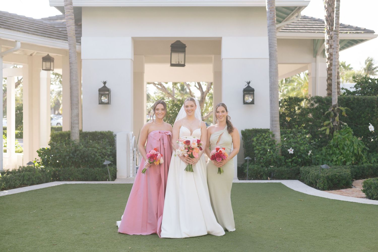 Elegant wedding party gathers on white columned porch with pink floral bouquets.