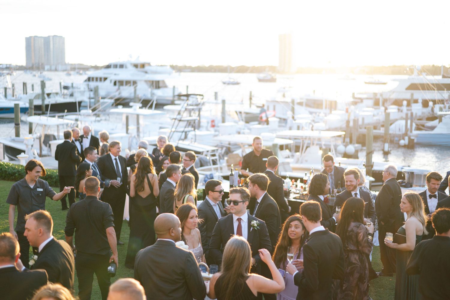 Wedding guests mingle at an outdoor marina reception during golden hour with boats in the background.